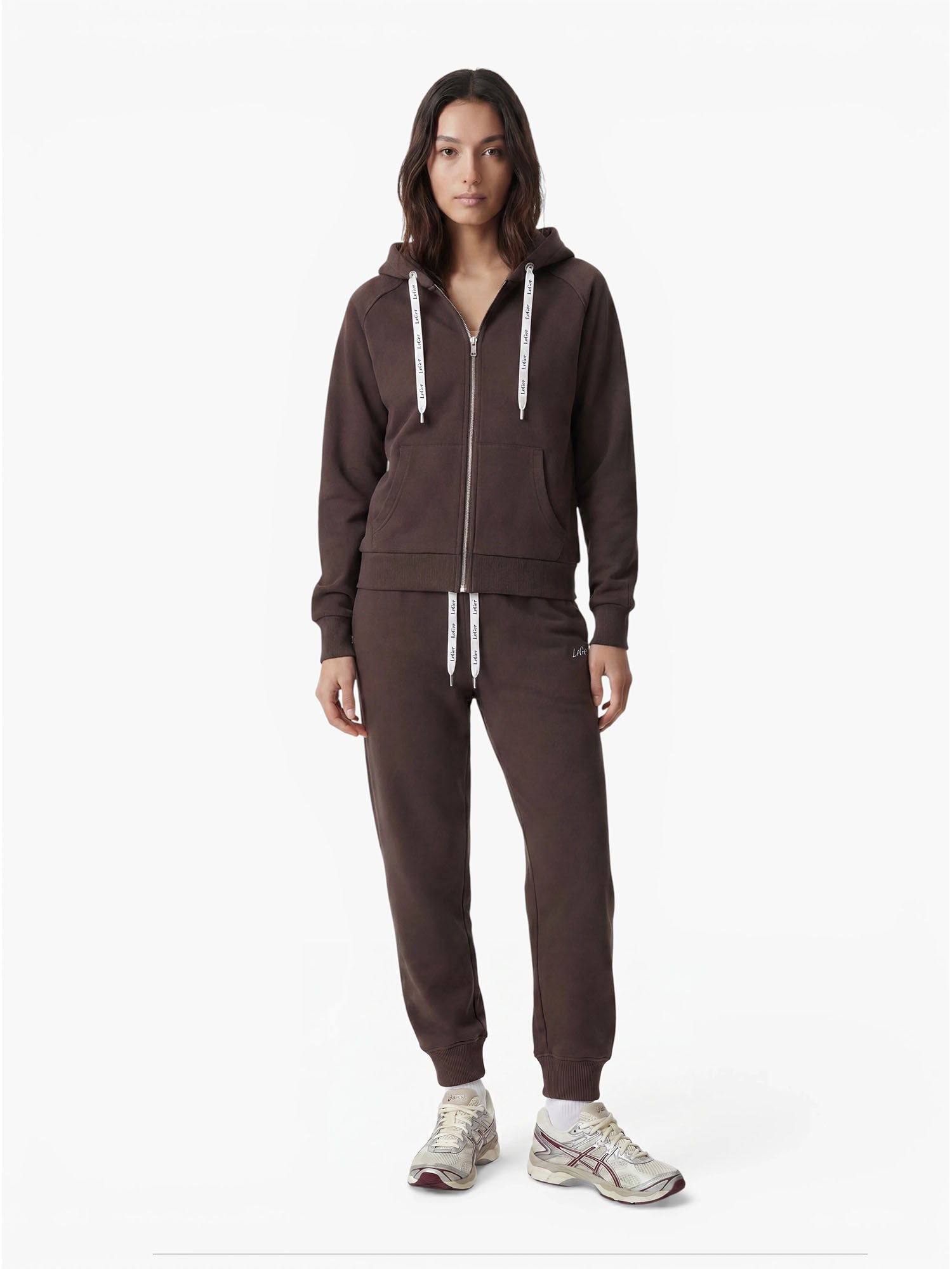 Eine Person steht mit dem Gesicht nach vorne vor einem schlichten weißen Hintergrund und trägt einen dunkelbraunen Kapuzenpulli mit Reißverschluss sowie eine Hose im Modell „Nela“ von LeGer by Lena Gercke mit taillierter Passform und weißen Kordeln, dazu hellgraue Sneaker.