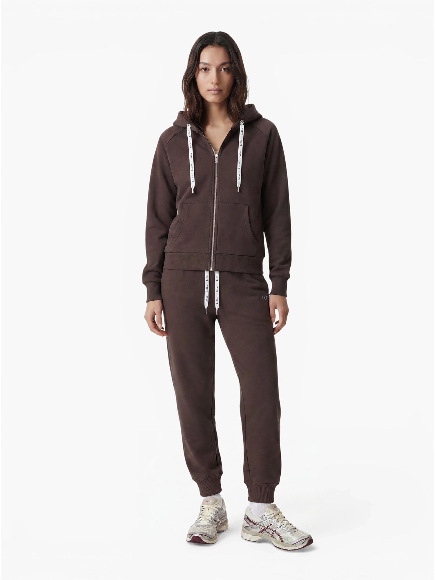 Eine Person steht mit dem Gesicht nach vorne, gekleidet in einen passenden dunkelbraunen Hoodie und ein Jogginghosen-Set der Marke „LeGer by Lena Gercke“ im Modell „Nela“ mit weißen Kordelzügen, dazu hellfarbige Sneaker, vor einem schlichten weißen Hintergrund.