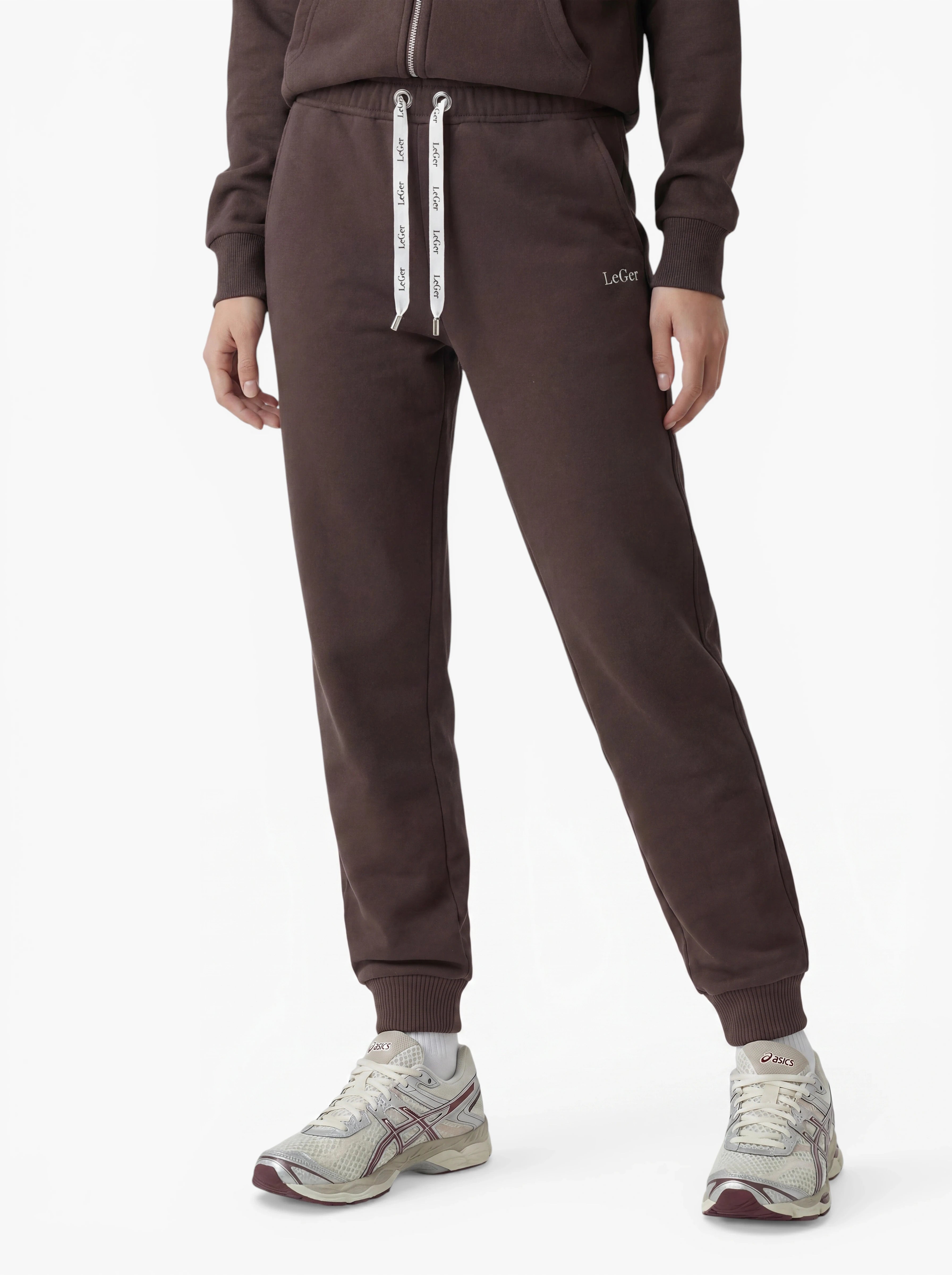 Klar! Hier ist eine überarbeitete Version deines Satzes unter Verwendung der bereitgestellten Produktdaten: Eine Person trägt die „Nela Pants“ von LeGer by Lena Gercke in Braun mit taillierter Passform und weißen Kordelzügen – gefertigt aus weichem Stretchstoff –, dazu einen passenden braunen Kapuzenpulli mit Reißverschluss und weiße Sneaker mit grauen und kastanienbraunen Akzenten, vor einem schlichten weißen Hintergrund.