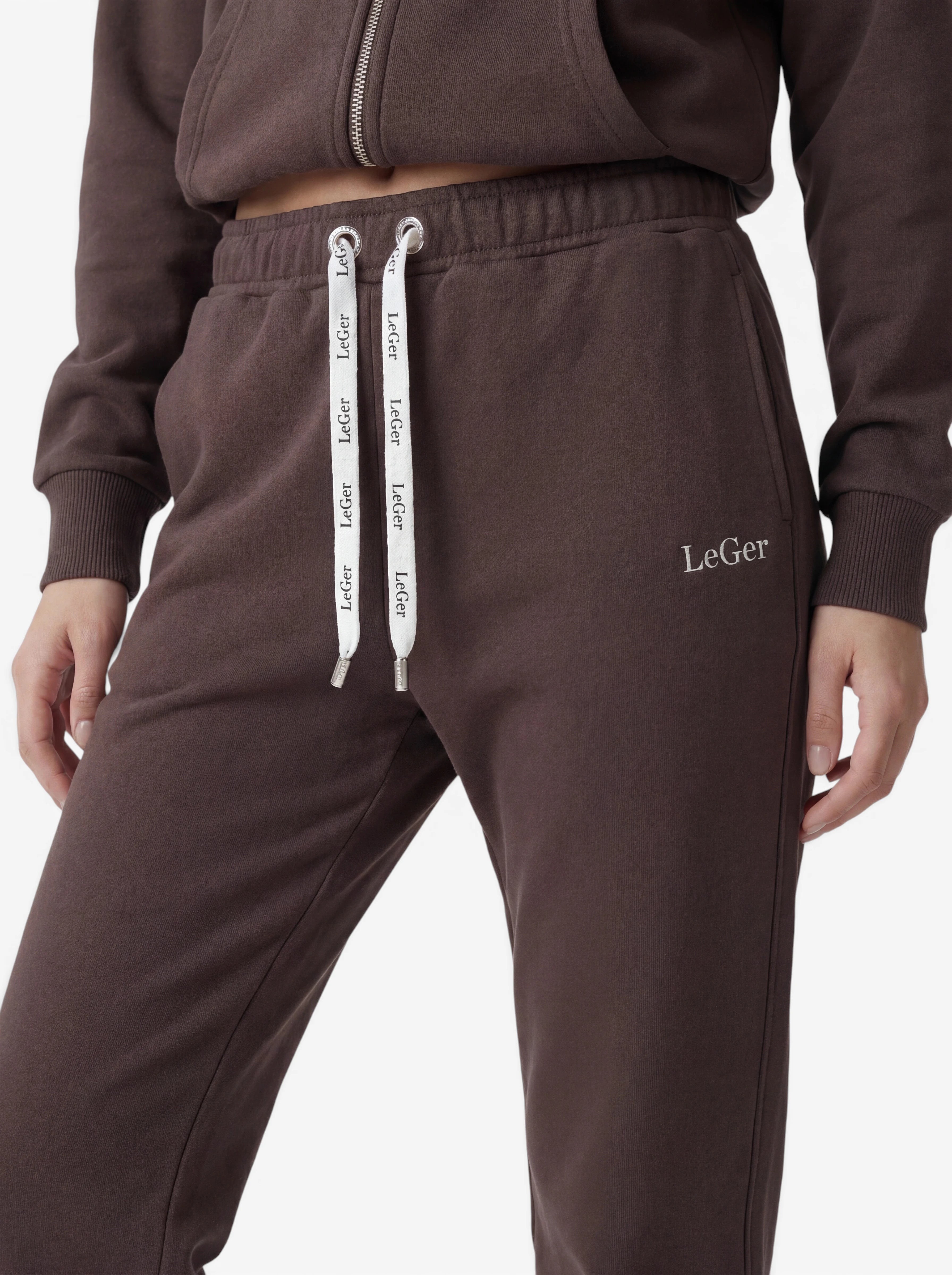 Zu sehen ist eine Person, die eine dunkelbraune „Nela“-Hose von LeGer by Lena Gercke trägt, kombiniert mit einem passenden Sweatshirt mit Reißverschluss. Die Jogginghose ist aus angenehm weichem Stretchstoff gefertigt und verfügt über eine taillierte Passform für eine vorteilhafte Silhouette. Weiße Kordeln mit dem Schriftzug „LeGer“ sowie das „LeGer“-Logo auf der linken Tasche sorgen für charakteristische Details. Die Hand der Person ruht an der Seite vor einem weißen Hintergrund.