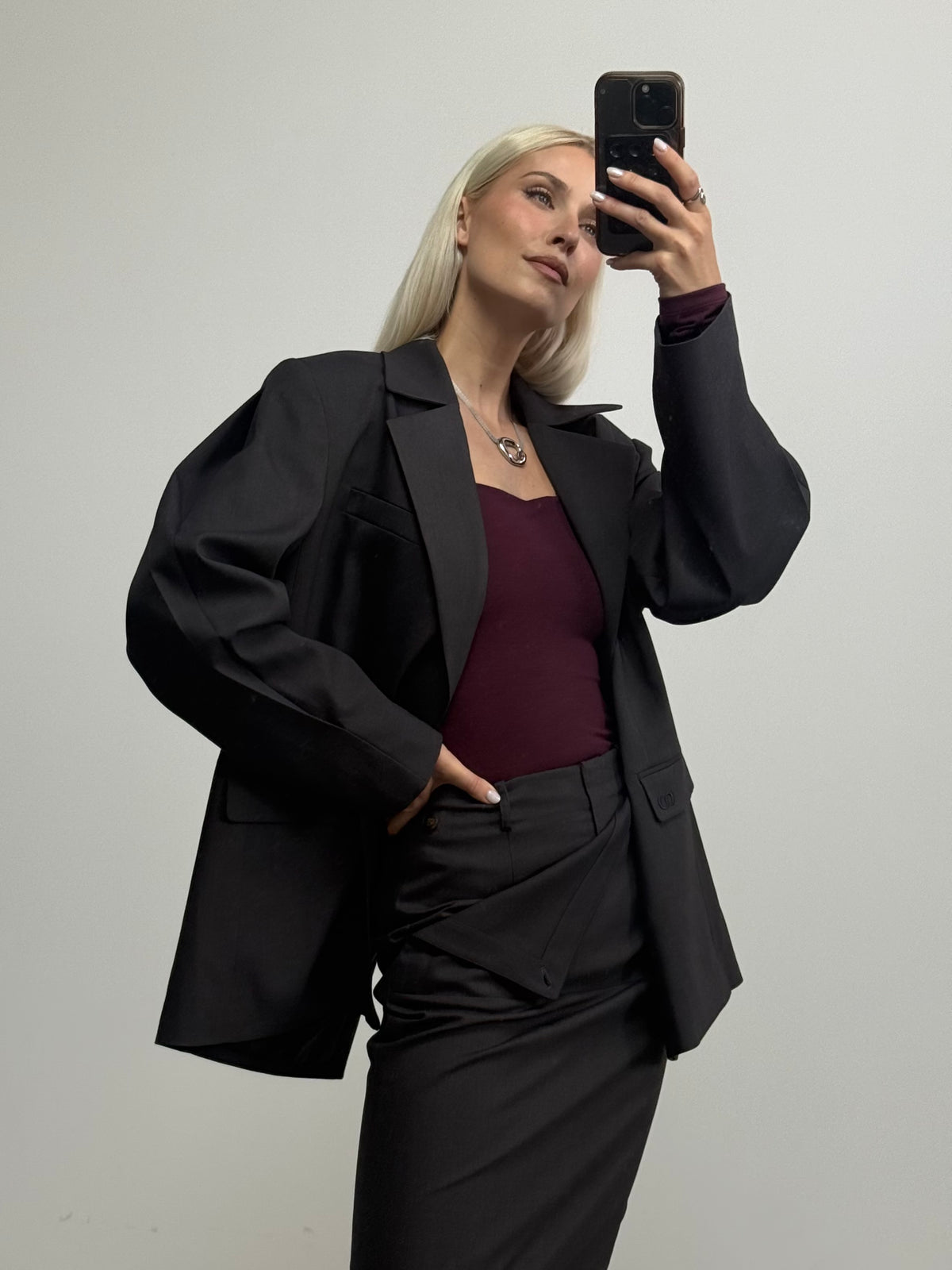 Eine Frau mit langen blonden Haaren trägt einen schwarzen Blazer und einen dazu passenden, eng anliegenden Rock, dazu das „Elorah“-Langarmshirt von LeGer by Lena Gercke mit Karree-Ausschnitt in Burgunderrot, während sie vor einem schlichten weißen Hintergrund für ein Spiegel-Selfie posiert.
