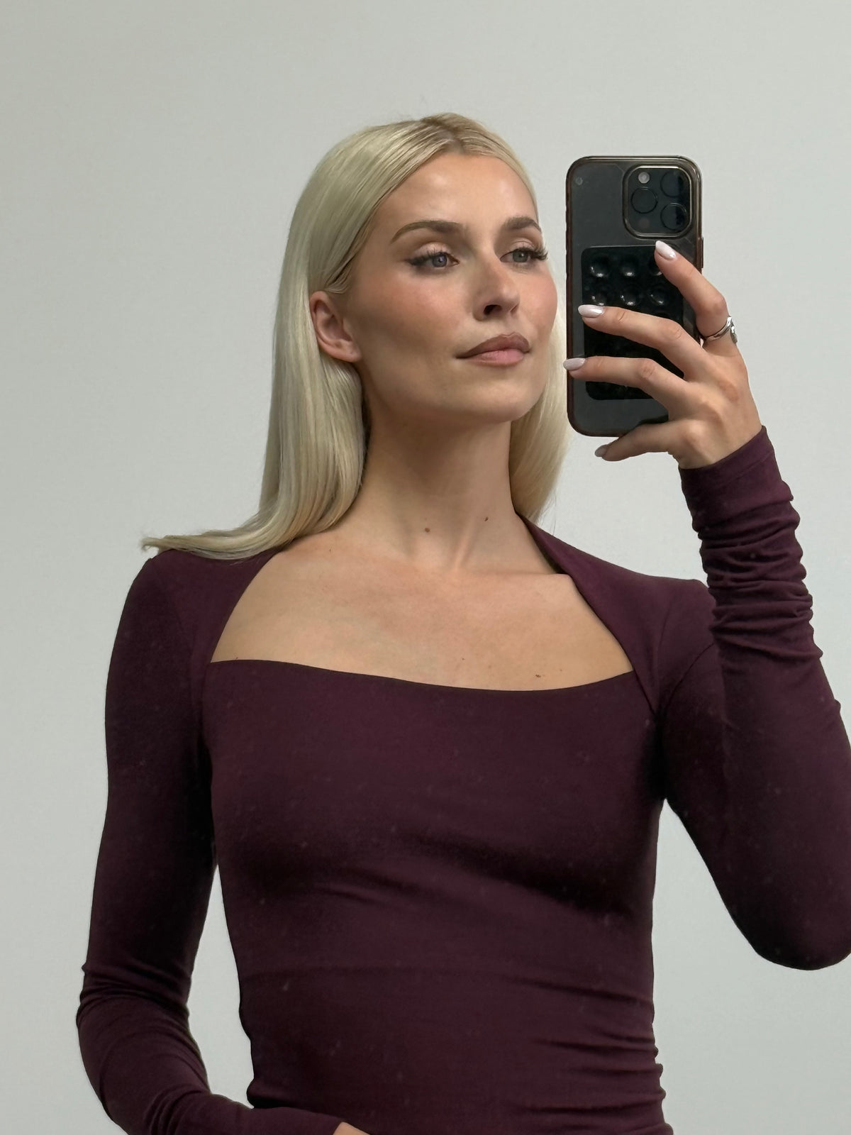 Eine Frau mit langen blonden Haaren, die ein dunkelviolettes „Elorah“-Langarmshirt von LeGer by Lena Gercke mit quadratischem Ausschnitt trägt, macht mit ihrem Smartphone ein Spiegel-Selfie. Der schmale Schnitt des Oberteils betont ihre Silhouette vor dem schlichten, hellen Hintergrund.