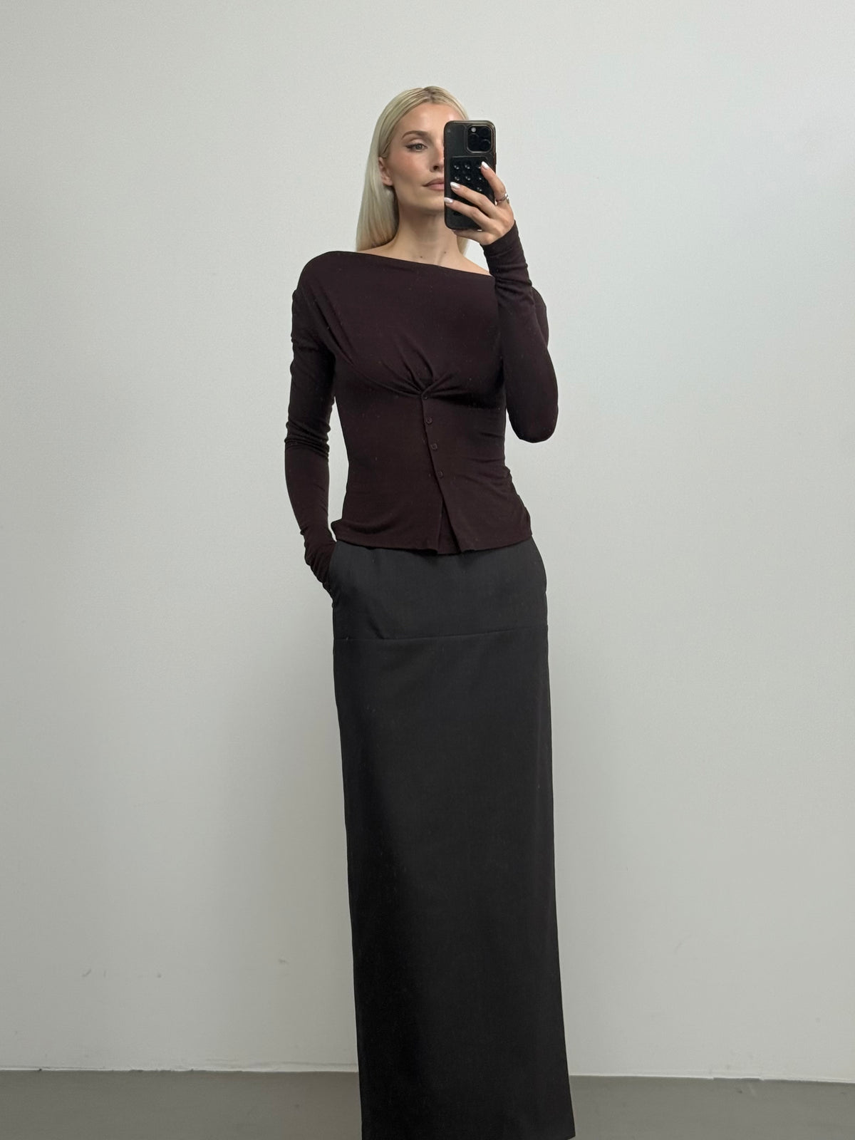 Eine Person steht in einem Innenraum vor einer schlichten weißen Wand. Sie trägt einen langen schwarzen Rock und das Langarmshirt „Regina“ von LeGer by Lena Gercke in Dunkelbraun mit einem stilvollen Wasserfallausschnitt und macht mit einer Hand in der Hosentasche ein Spiegel-Selfie.