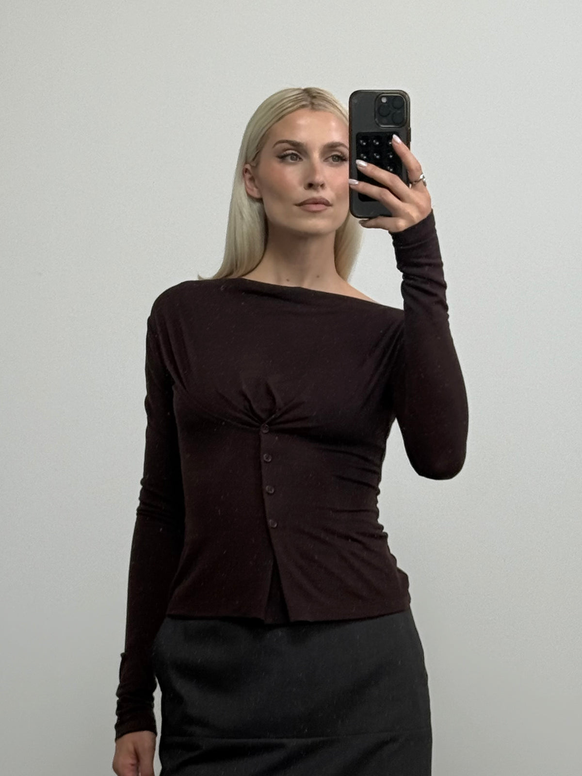 Eine Frau mit langen blonden Haaren macht ein Spiegel-Selfie. Sie trägt das Langarmshirt „Regina“ von LeGer by Lena Gercke in Dunkelbraun mit Knopfleiste auf der Vorderseite, dazu einen dunklen Rock, und steht vor einem schlichten hellen Hintergrund.