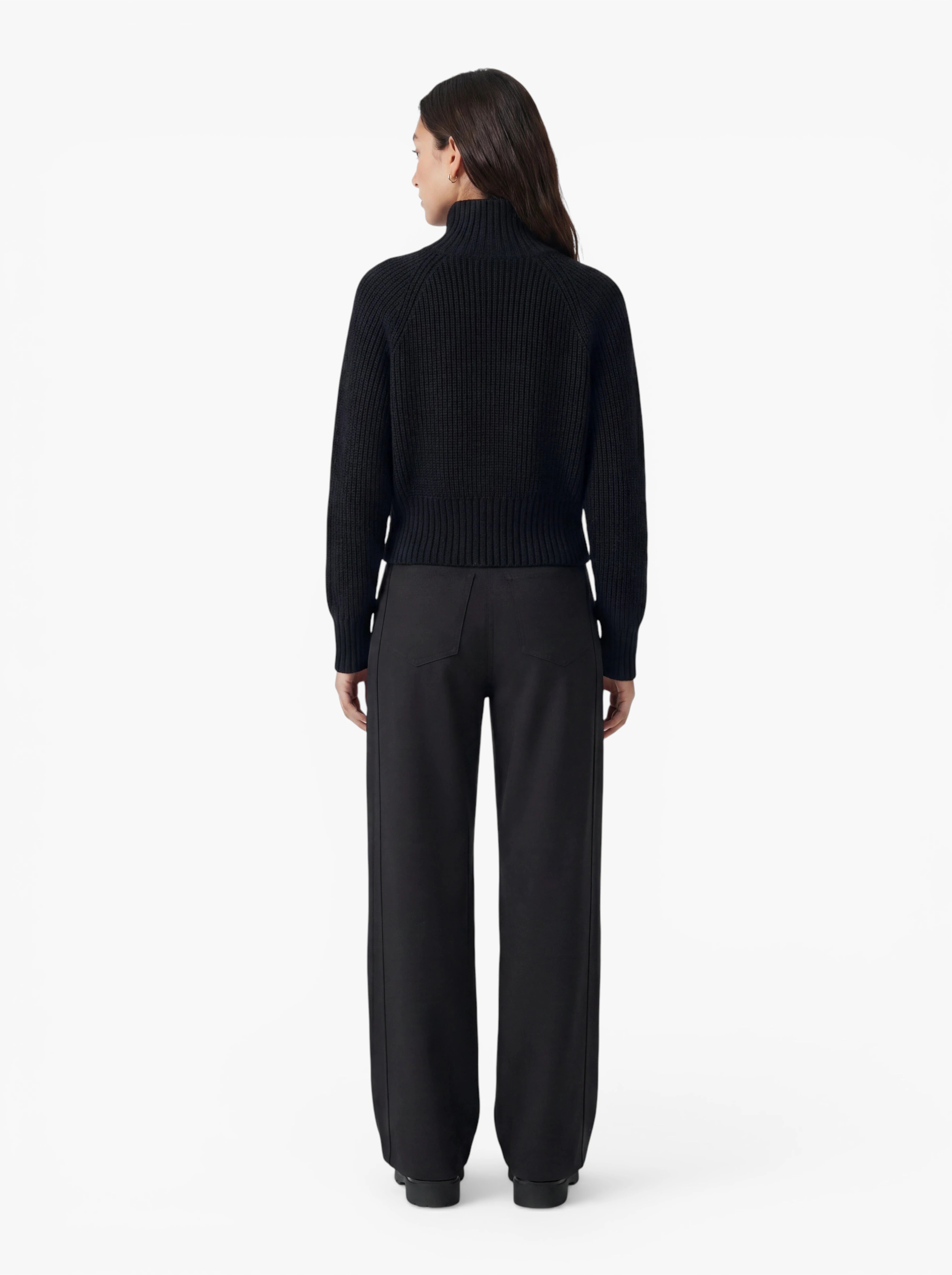 Eine Frau steht mit dem Rücken zur Kamera vor einem schlichten weißen Hintergrund und trägt den schwarzen Pullover „Nala“ von LeGer by Lena Gercke mit einer entspannten Passform sowie eine schwarze Hose mit weitem Bein.