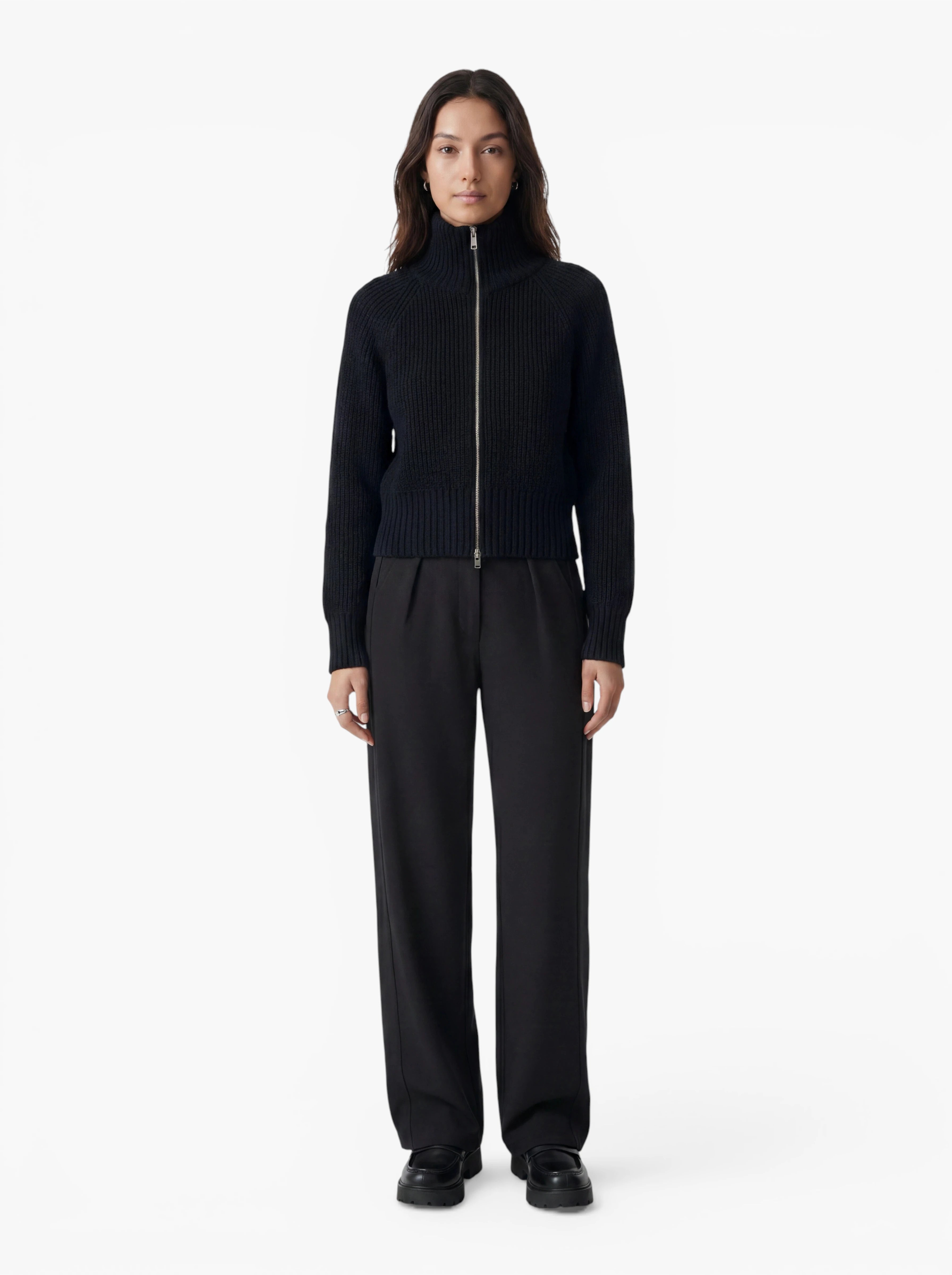 Vor einem schlichten weißen Hintergrund steht eine Person mit dem Gesicht nach vorne, bekleidet mit dem Pullover „Nala“ von LeGer by Lena Gercke – einem schwarzen Pullover mit Reißverschluss und entspannter Passform –, einer lockeren schwarzen Hose und schwarzen Schuhen.
