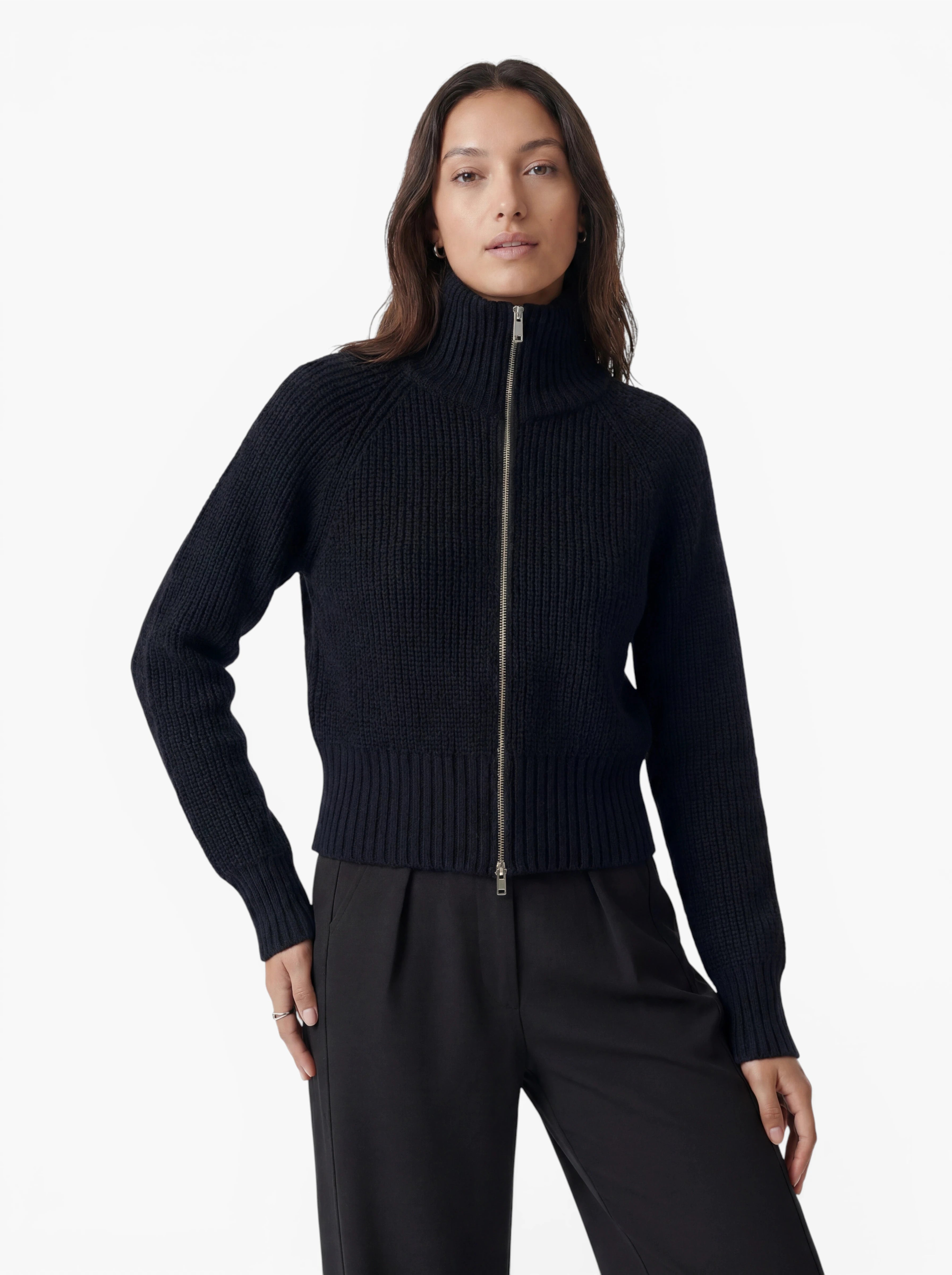 Eine Frau, die den Pullover „Nala“ aus der Kollektion „LeGer by Lena Gercke“ trägt – einen schwarzen, gerippten Pullover mit Reißverschluss und entspannter Passform –, steht zusammen mit einer schwarzen Hose vor einem schlichten weißen Hintergrund und blickt mit einer Hand in die Hüfte gestützt nach vorne. Dieser Strickpullover vereint Komfort und Eleganz.
