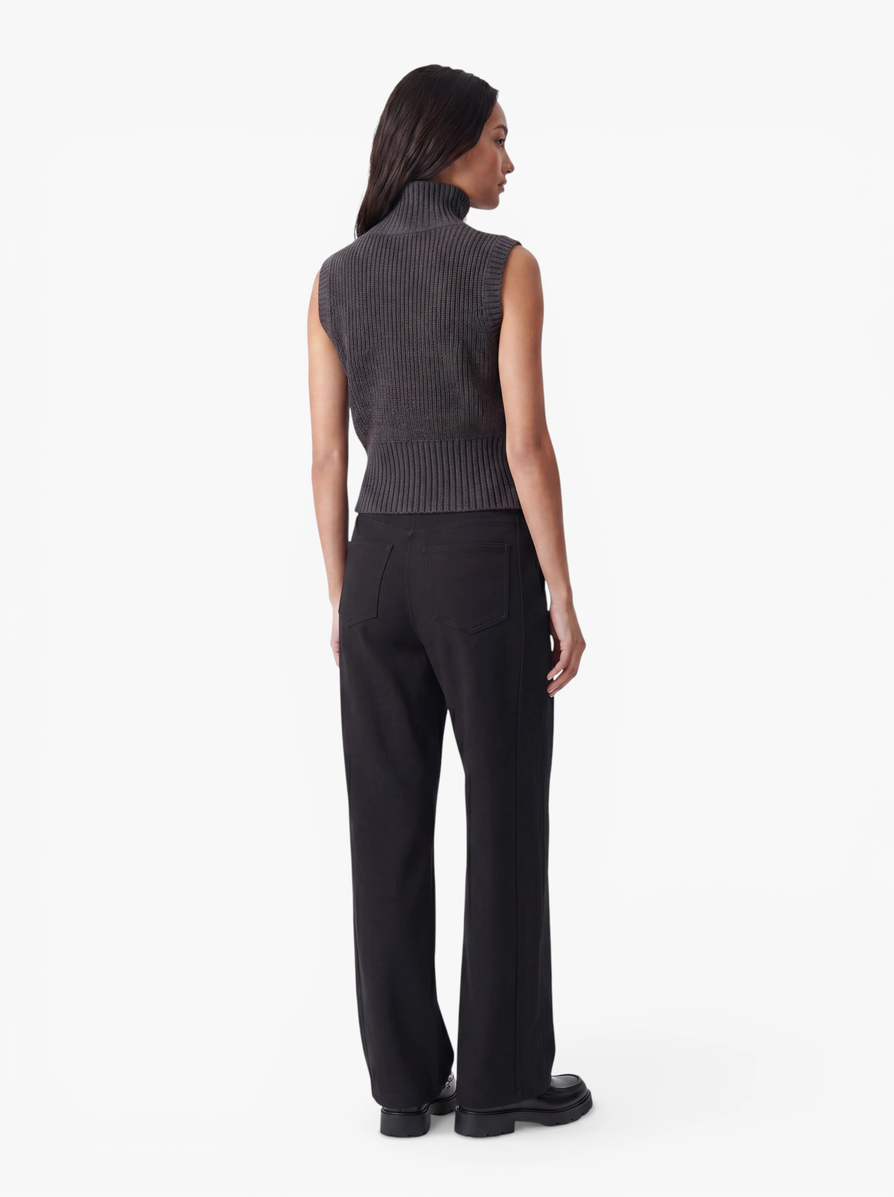 Eine Person steht mit dem Rücken zur Kamera und trägt die Weste „Betsy“ von LeGer by Lena Gercke – einen ärmellosen grauen Rollkragenpullover in minimalistischem Design –, dazu schwarze Hosen und schwarze Schuhe, vor einem schlichten weißen Hintergrund.