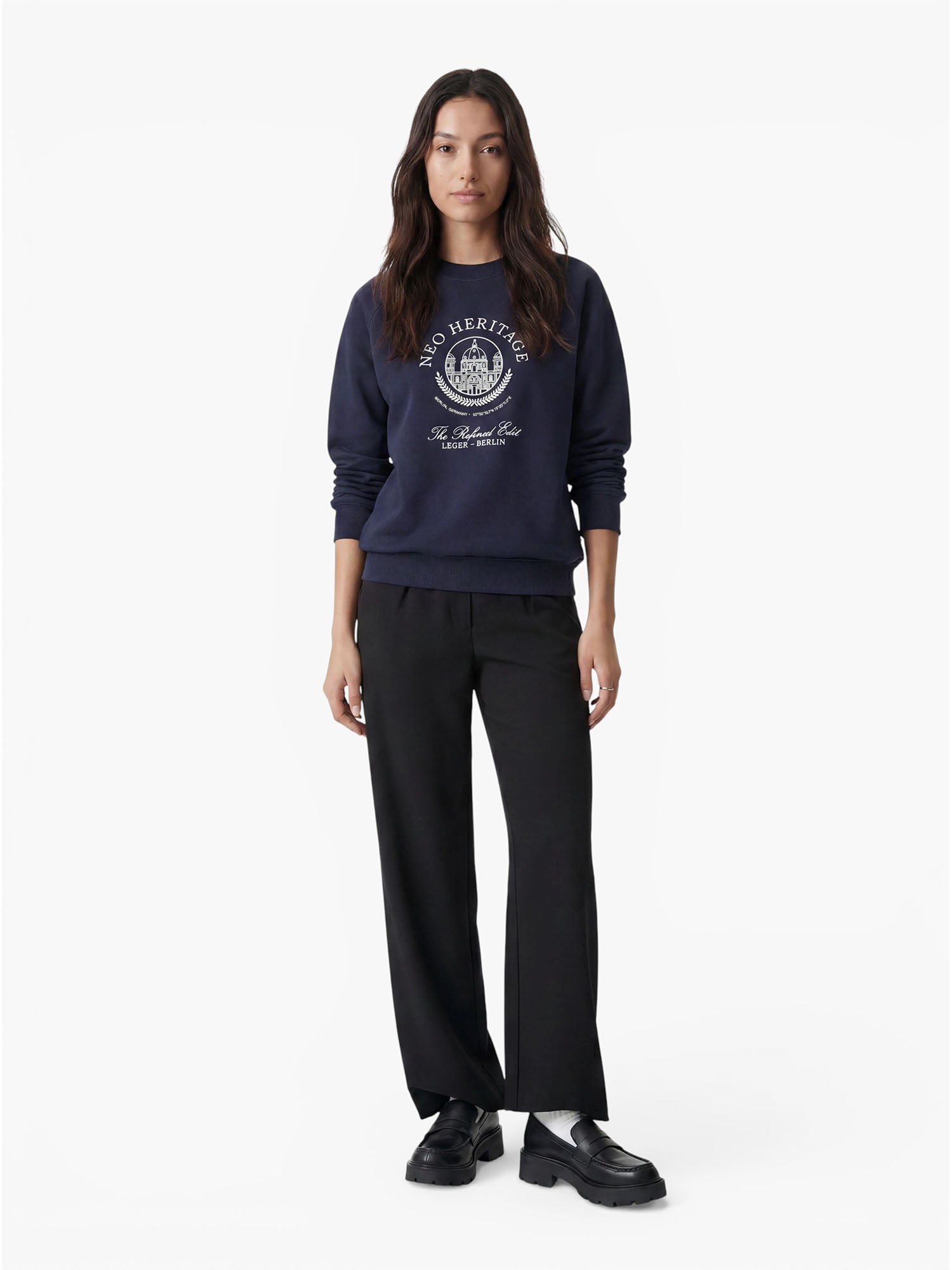 Eine Frau mit langen, dunklen Haaren steht mit dem Blick nach vorne und trägt den marineblauen „Bettina“-Pullover von LeGer by Lena Gercke in lockerer Passform mit weißer Aufschrift, dazu eine schwarze Hose und schwarze Schuhe – ein lässiger Look vor einem schlichten weißen Hintergrund.