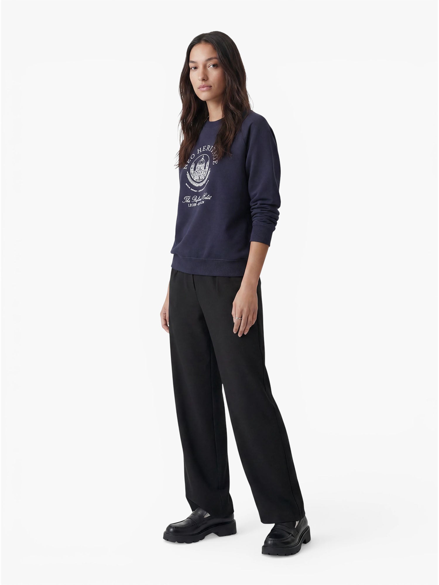 Eine Frau steht mit dem Blick nach vorne und trägt den Pullover „Bettina“ von LeGer by Lena Gercke in Marineblau mit weißem Aufdruck, kombiniert mit einer schwarzen Hose in lockerer Passform und schwarzen Plateauschuhen vor einem schlichten weißen Hintergrund – ein perfekter Look für den beliebten Casual-Mode-Stil.
