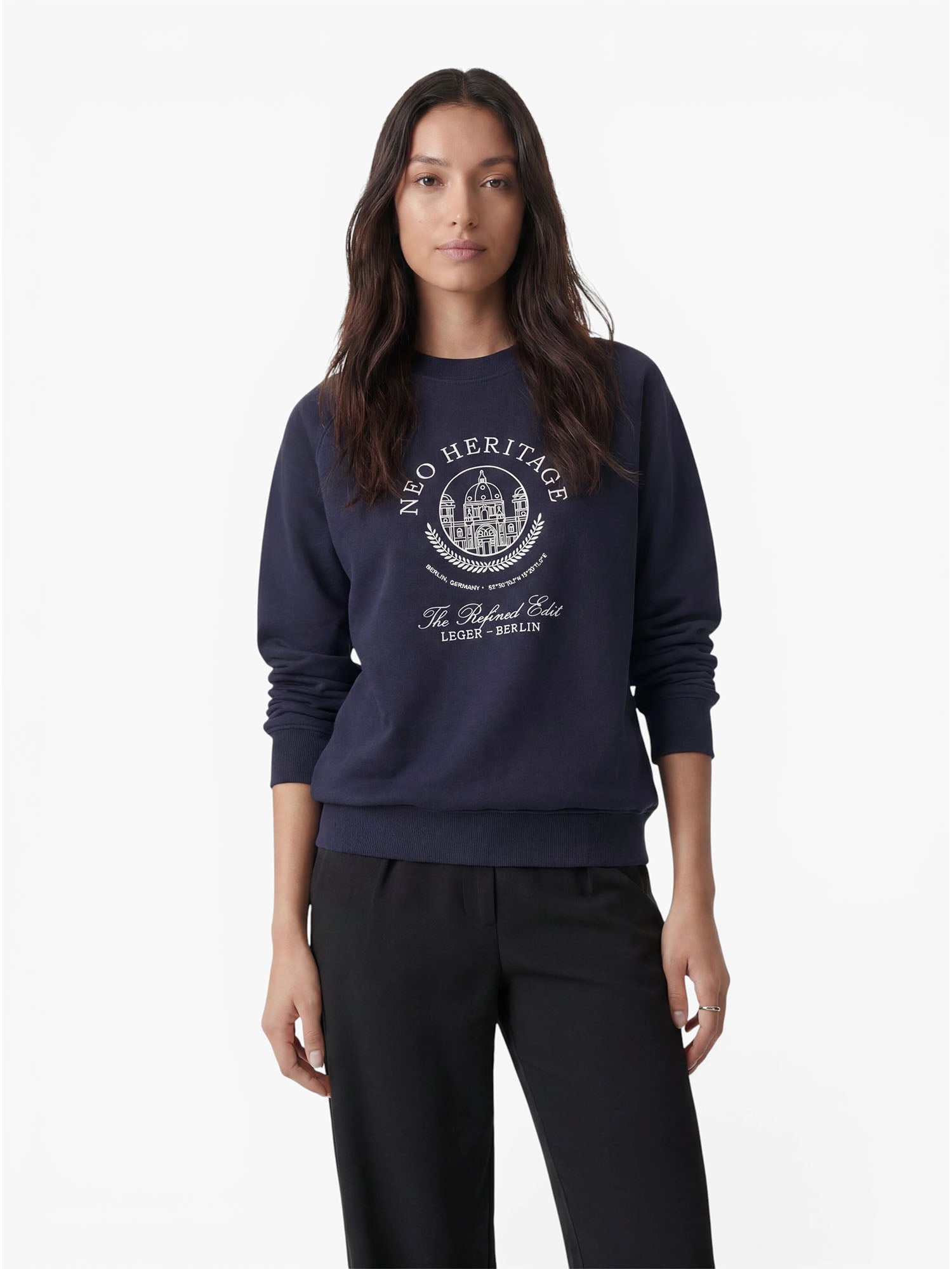 Eine Frau mit langen, dunklen Haaren trägt den Pullover „Bettina“ von LeGer by Lena Gercke in Marineblau, der sich durch ein weißes, rundes Logo und einen Schriftzug sowie eine lockere Passform auszeichnet. Sie kombiniert ihn mit einer schwarzen Hose und steht vor einem schlichten weißen Hintergrund – ein mühelos lässiger Look.