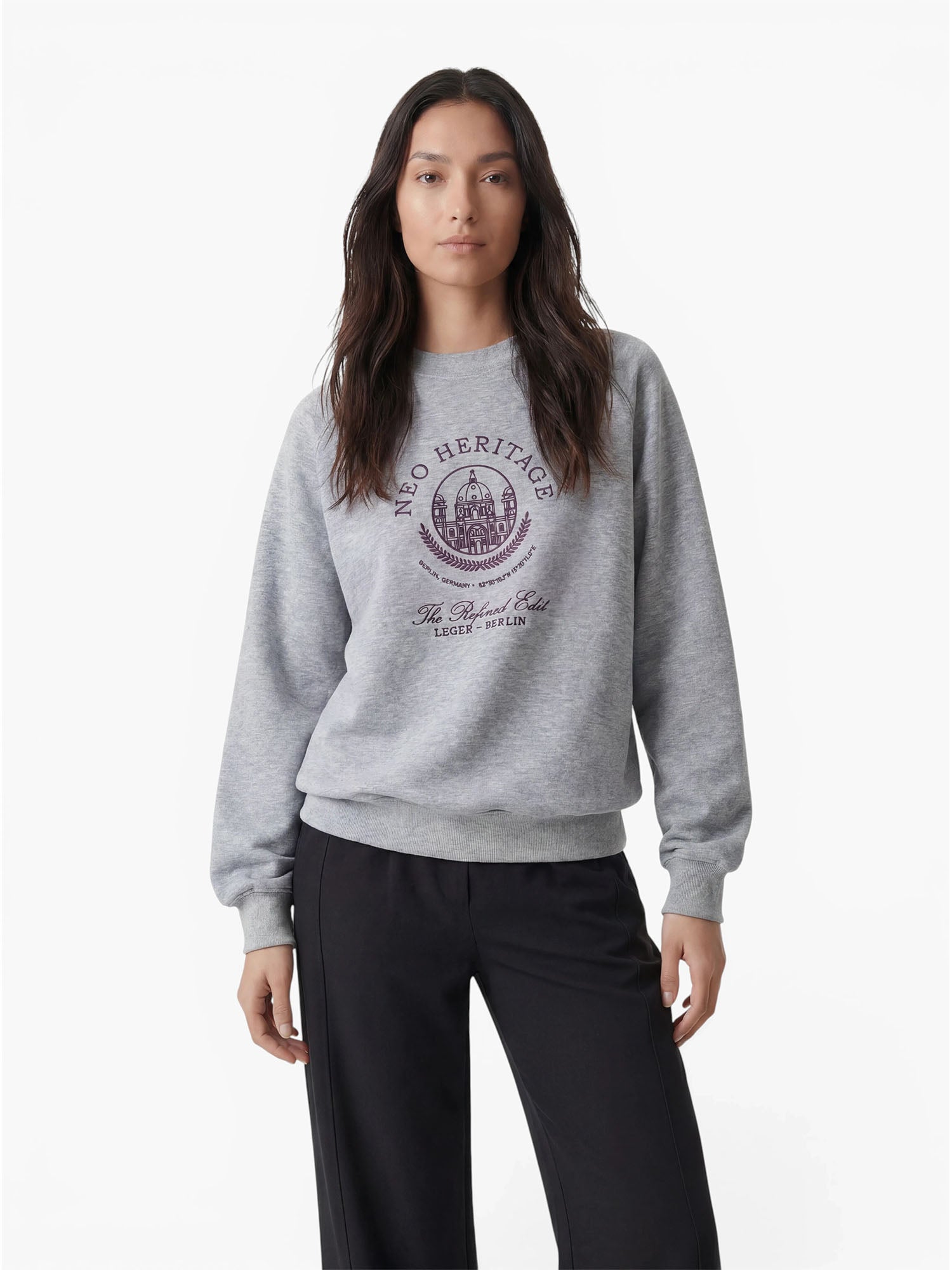Eine Person mit langen, dunklen Haaren trägt den Pullover „Bettina“ von LeGer by Lena Gercke, ein bequemes graues Sweatshirt mit einem violetten Wappen und einem Schriftzug, dazu eine schwarze Hose; sie steht vor einem schlichten weißen Hintergrund.