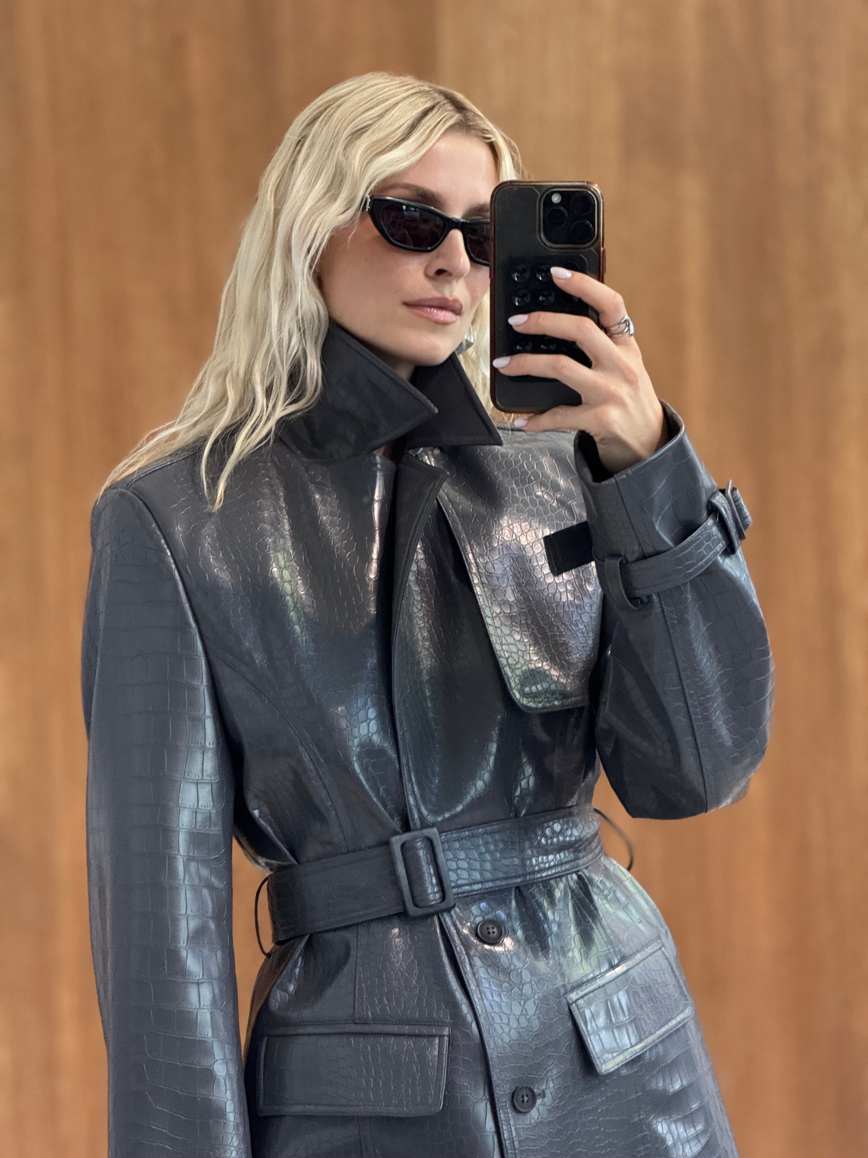 Eine Person mit langen blonden Haaren, die eine Sonnenbrille trägt und den glänzenden, strukturierten „Natascha“-Mantel von LeGer by Lena Gercke, macht mit ihrem Handy ein Spiegel-Selfie. Im Hintergrund ist eine schlichte Wand in warmen Farbtönen zu sehen.