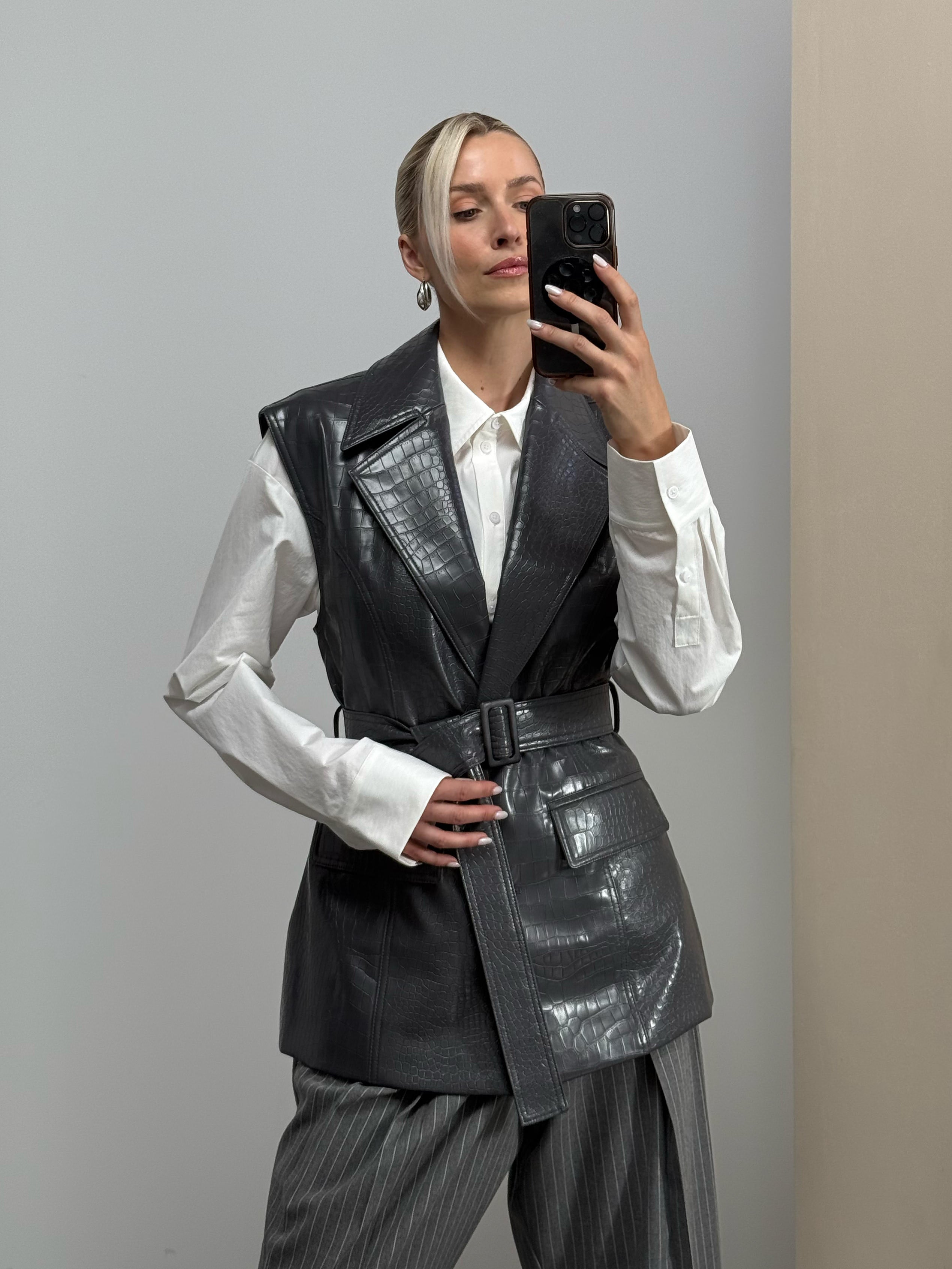 Eine Person, die ein Spiegel-Selfie macht. Sie trägt ein weißes Hemd, die Weste „Hanne“ von LeGer by Lena Gercke – eine elegante, moderne, mittellange Weste aus schwarzem Leder, die hohen Tragekomfort bietet – sowie eine graue Nadelstreifenhose und steht vor einer schlichten, neutralen Wand.
