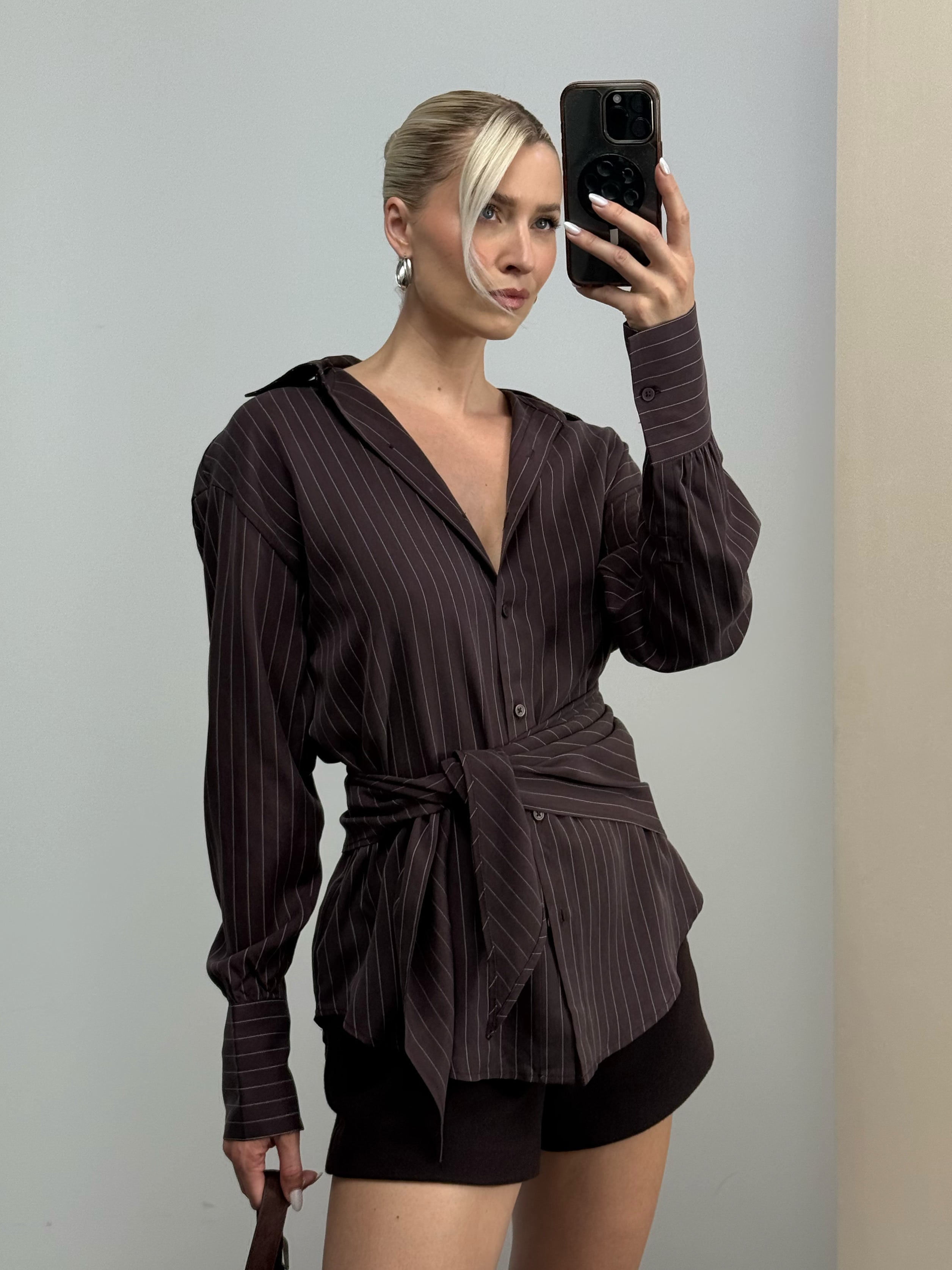 Eine Frau mit blonden Haaren, die zu einem tief sitzenden Dutt zusammengebunden sind, macht ein Spiegel-Selfie. Sie trägt die Bluse „Beyond“ von LeGer by Lena Gercke – eine locker sitzende, dunkel gestreifte Damen-Businessbluse mit einem Bindegürtel in der Taille –, kombiniert mit schwarzen Shorts und Ohrringen, vor einem hellen Hintergrund.
