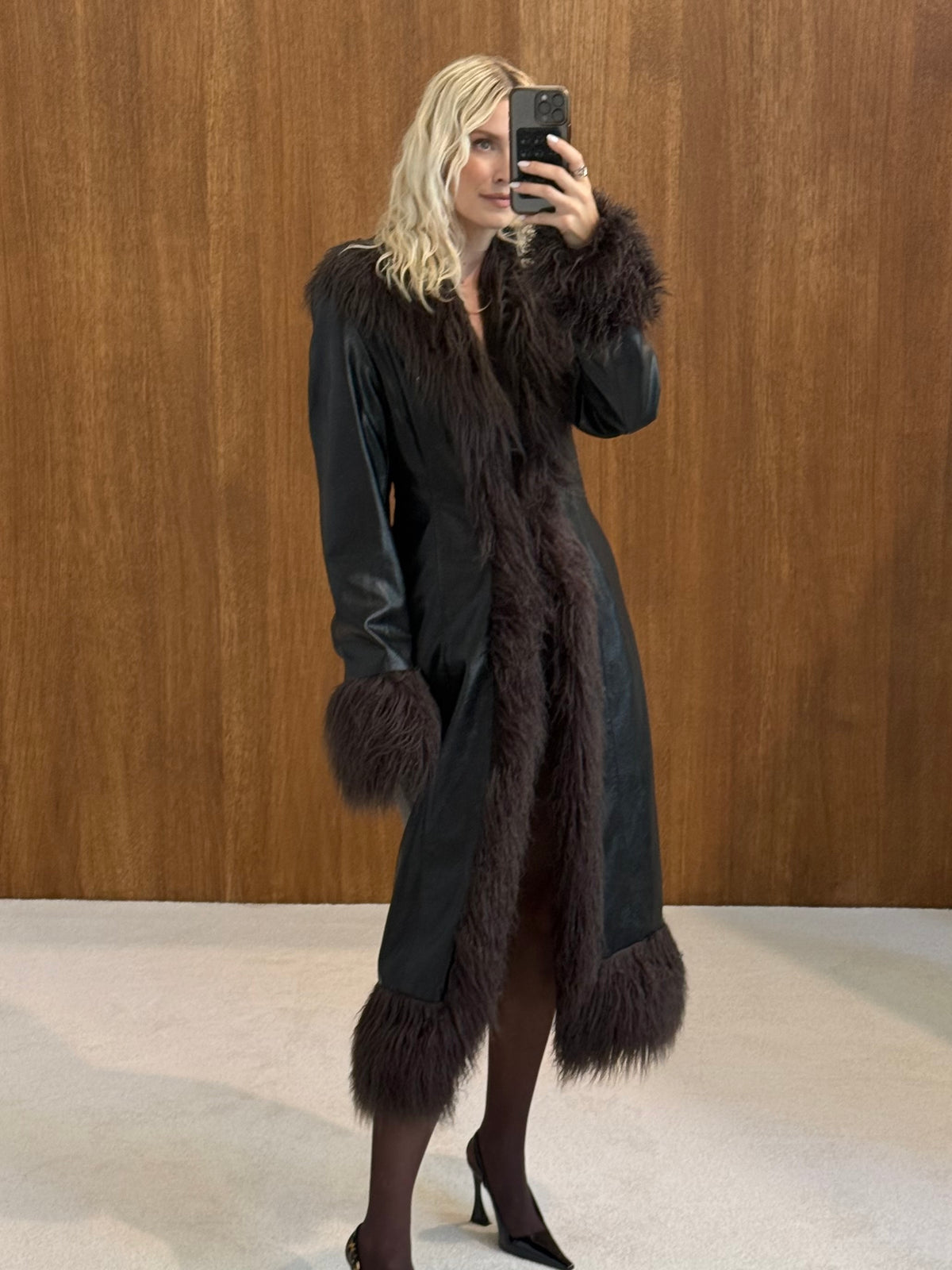 Eine Frau mit welligem blondem Haar trägt den taillierten Damenmantel „Teena“ von LeGer by Lena Gercke mit dunklem Kunstfellbesatz, dazu schwarze Pumps und dunkle Strumpfhosen, während sie vor einer Holzwand ein Selfie macht.
