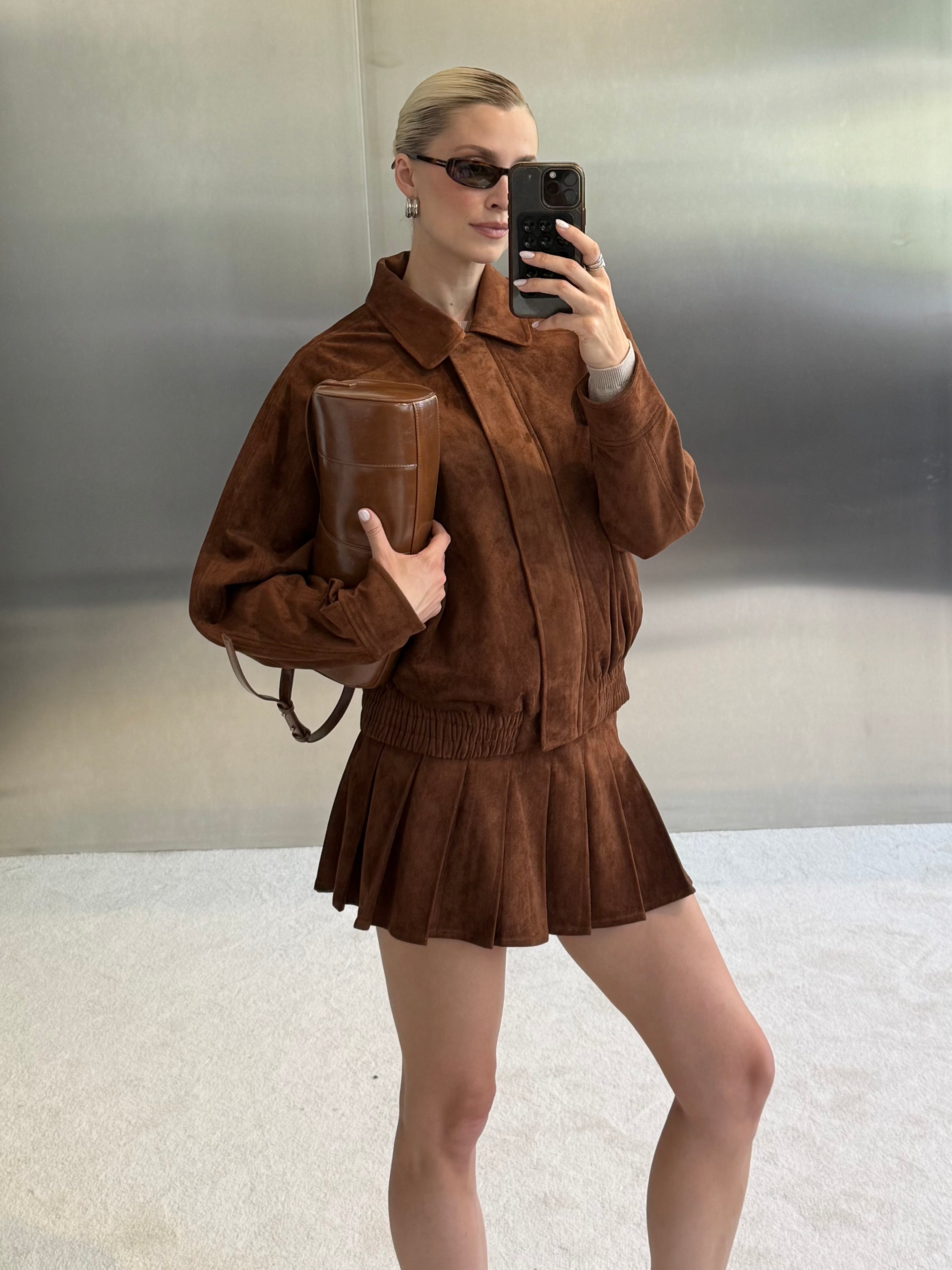 Eine Person mit Sonnenbrille macht ein Spiegel-Selfie: Sie trägt eine braune Wildlederjacke und den stilvollen Rock „Armine“ von LeGer by Lena Gercke und hält eine braune Clutch in der Hand. Der Hintergrund ist ein moderner, minimalistischer Raum mit metallischen Wänden.