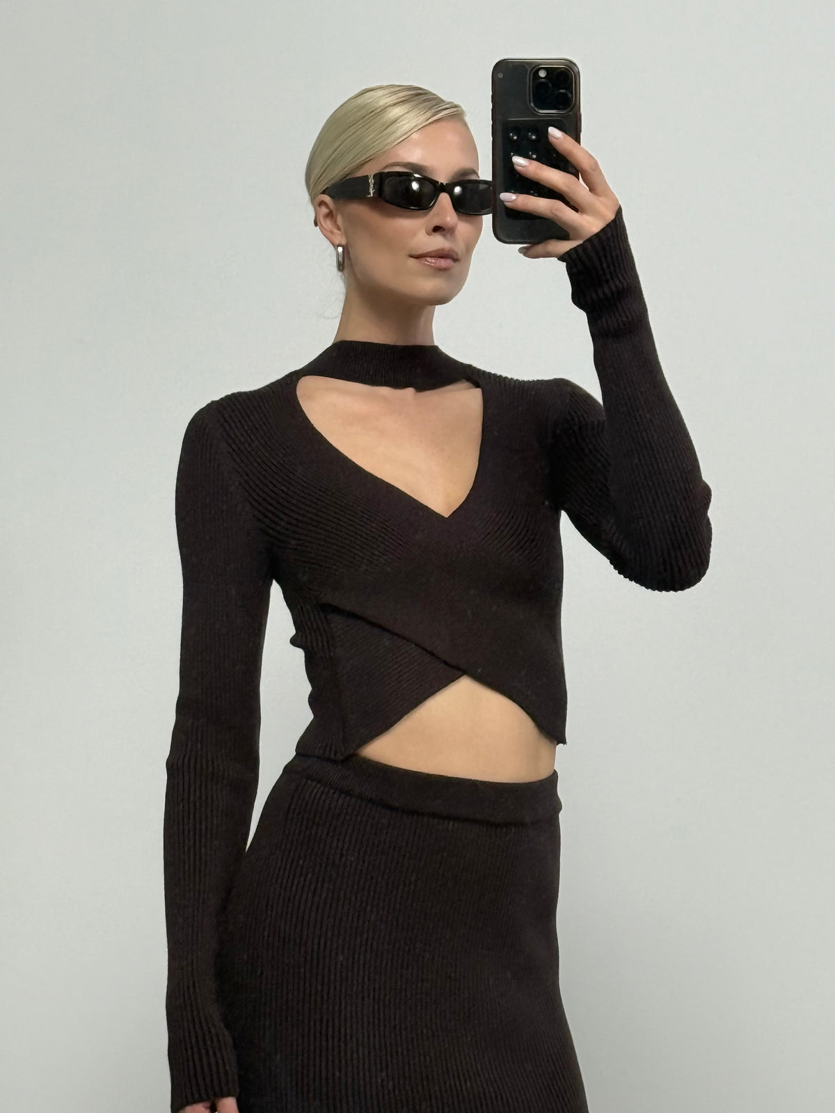 Eine Person mit schwarzer Sonnenbrille und einem stilvollen schwarzen Strickoutfit, bestehend aus dem Rock „Juliena“ von LeGer by Lena Gercke im hoch taillierten Bleistiftrock-Design, macht mit ihrem Handy vor einem schlichten, hellen Hintergrund ein Selfie.