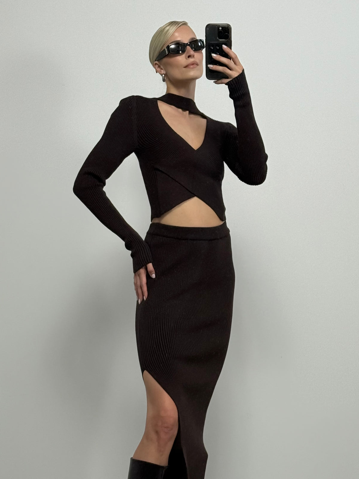 Eine Person trägt den stilvollen Pullover „Juliena“ von LeGer by Lena Gercke in Dunkelbraun mit V-Ausschnitt und langen Ärmeln, dazu einen Rock mit hohem Schlitz, und posiert für ein Spiegel-Selfie. Sie hat kurzes blondes Haar, trägt eine Sonnenbrille und Creolen.