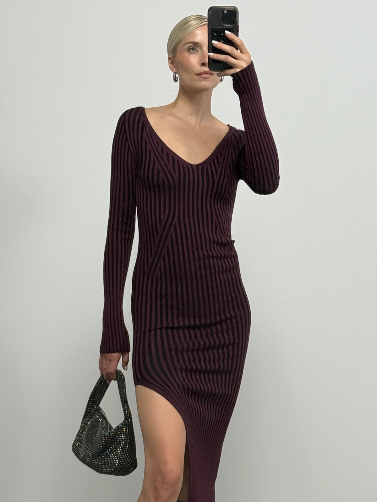Eine Frau trägt das Kleid „Juliena“ von LeGer by Lena Gercke – ein langärmeliges, gestreiftes, burgunderrotes Midikleid mit V-Ausschnitt und hohem Schlitz –, während sie eine kleine schwarze Handtasche in der Hand hält und vor einem schlichten weißen Hintergrund ein Spiegel-Selfie macht.