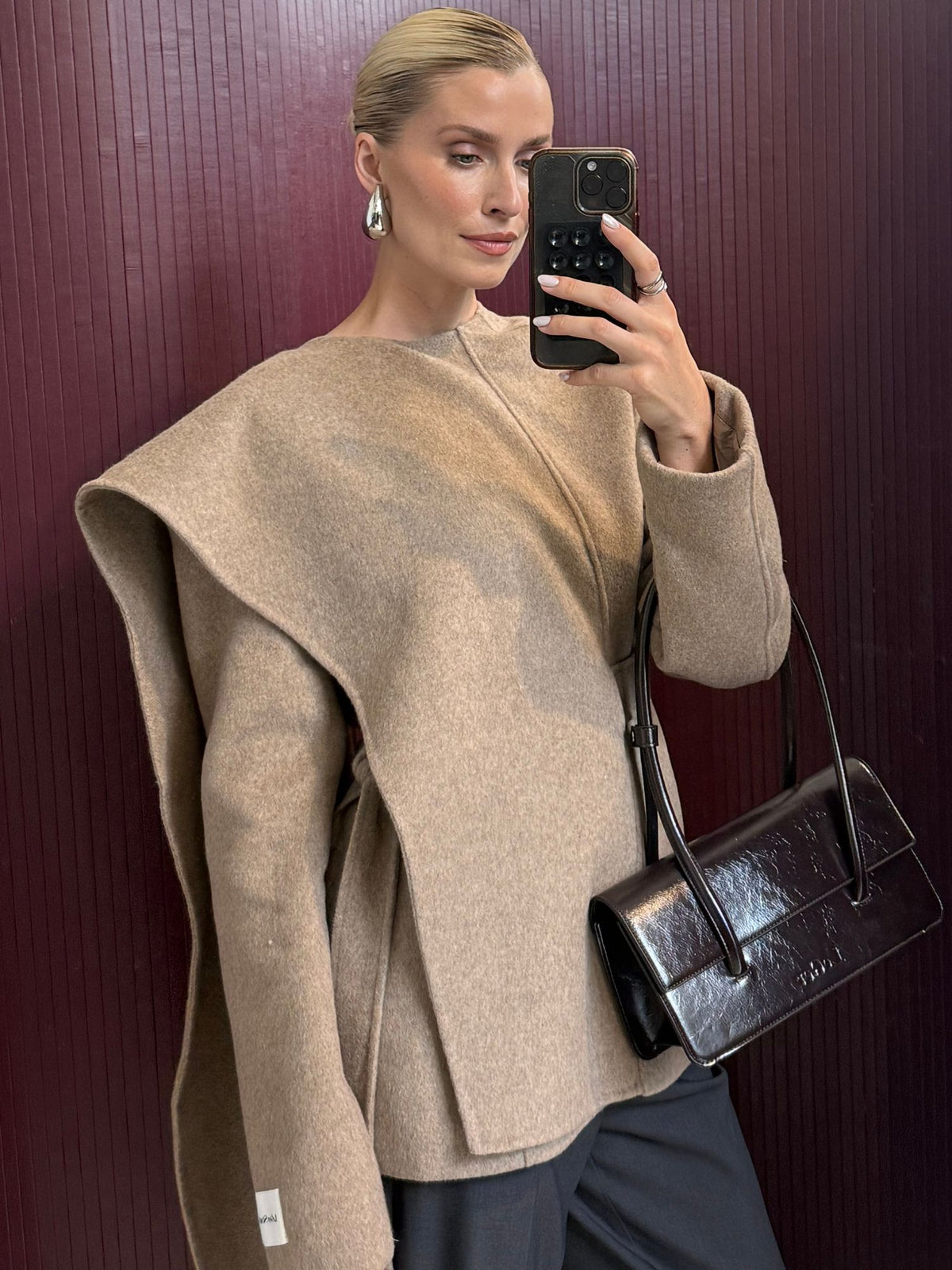 Eine Frau macht ein Spiegel-Selfie in der Jacke „Elva“ von LeGer by Lena Gercke – einem beigen, skulpturalen Modell in asymmetrischer Cape-Form mit übertriebenen Schultern –, kombiniert mit einer schwarzen Hose, silbernen Ohrringen und einer glänzenden schwarzen Handtasche. Die dunkelroten vertikalen Streifen im Hintergrund bilden einen auffälligen Kontrast, der ihr elegantes LeGer-Outfit besonders zur Geltung bringt.