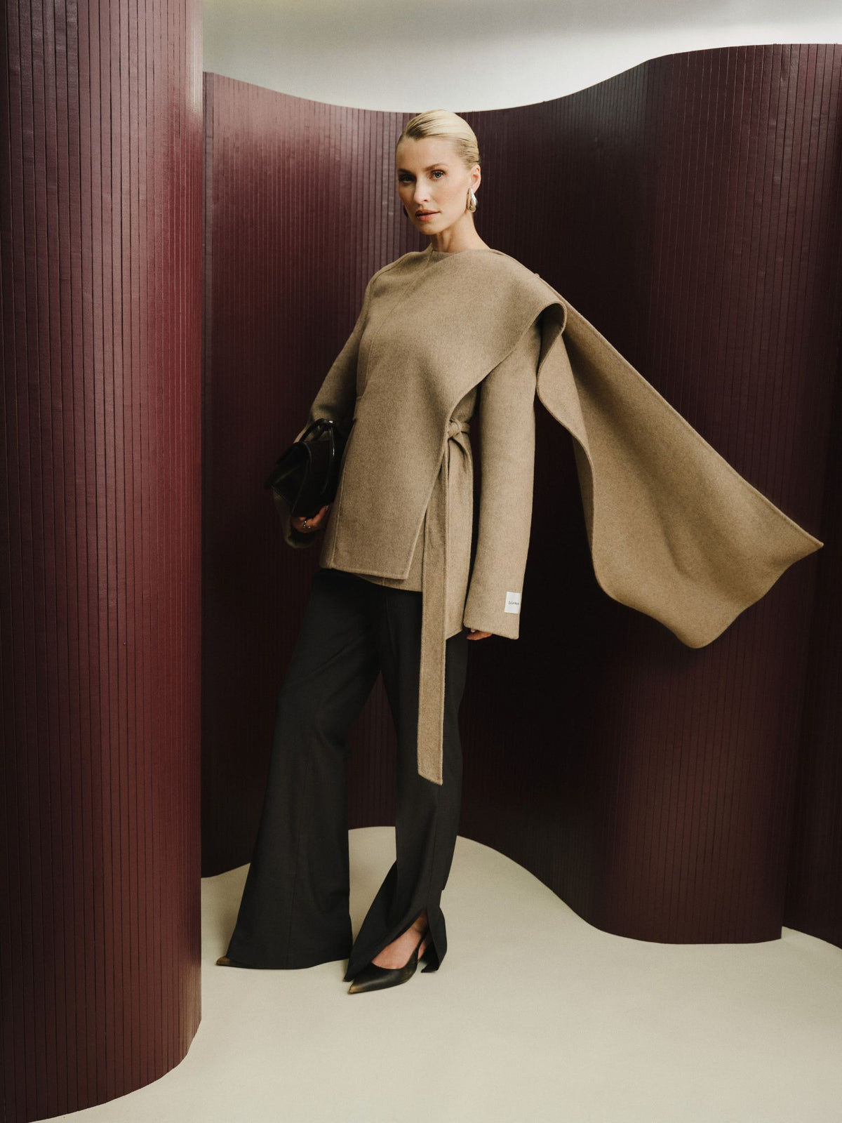 Eine Frau steht vor geschwungenen, burgunderroten Paneelen und trägt die Jacke „Elva“ von LeGer by Lena Gercke – ein beiges, asymmetrisches Oberteil in Cape-Form mit drapiertem Ärmel –, dazu eine schwarze Hose mit weitem Bein, schwarze Pumps und eine schwarze Clutch.