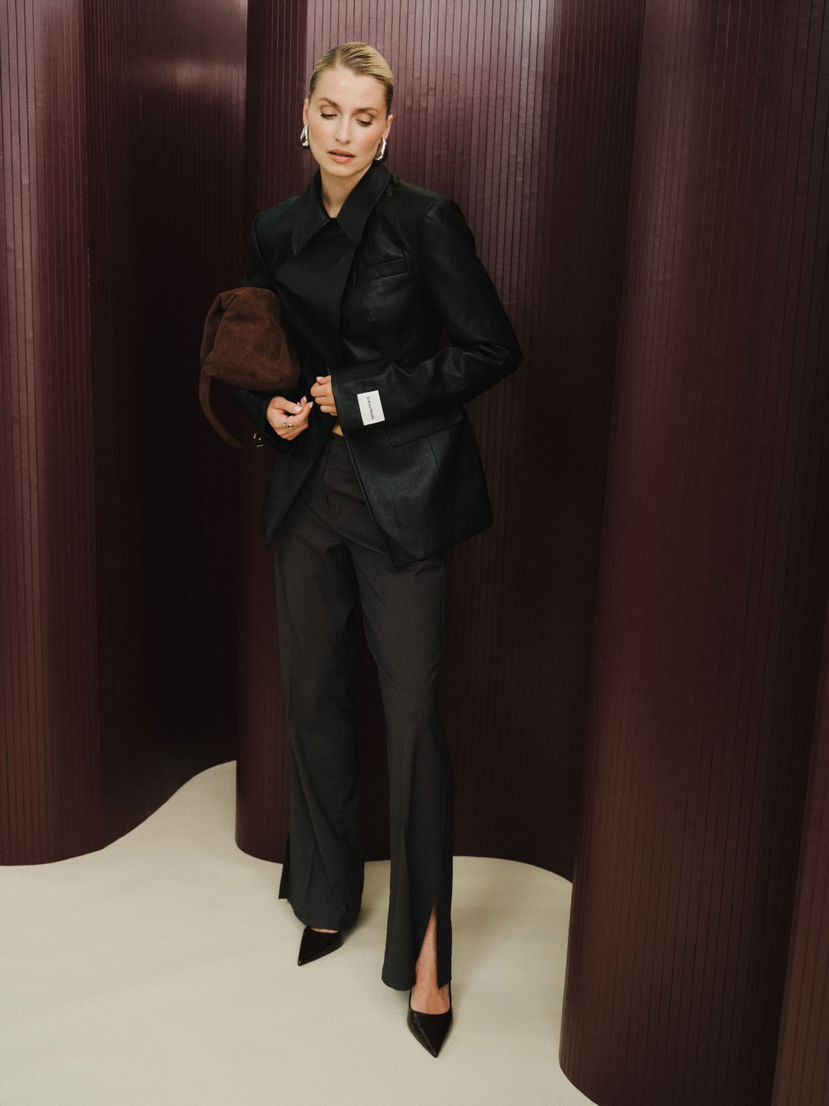 Eine Person, die den Blazer „Orelia“ von LeGer by Lena Gercke trägt – einen taillierten, schmal geschnittenen schwarzen Blazer in Lederoptik –, dazu eine dunkle Hose mit gespaltenem Saum und spitze Schuhe, steht vor hohen, burgunderroten, strukturierten Paneelen und hält eine braune Tasche unter dem Arm.