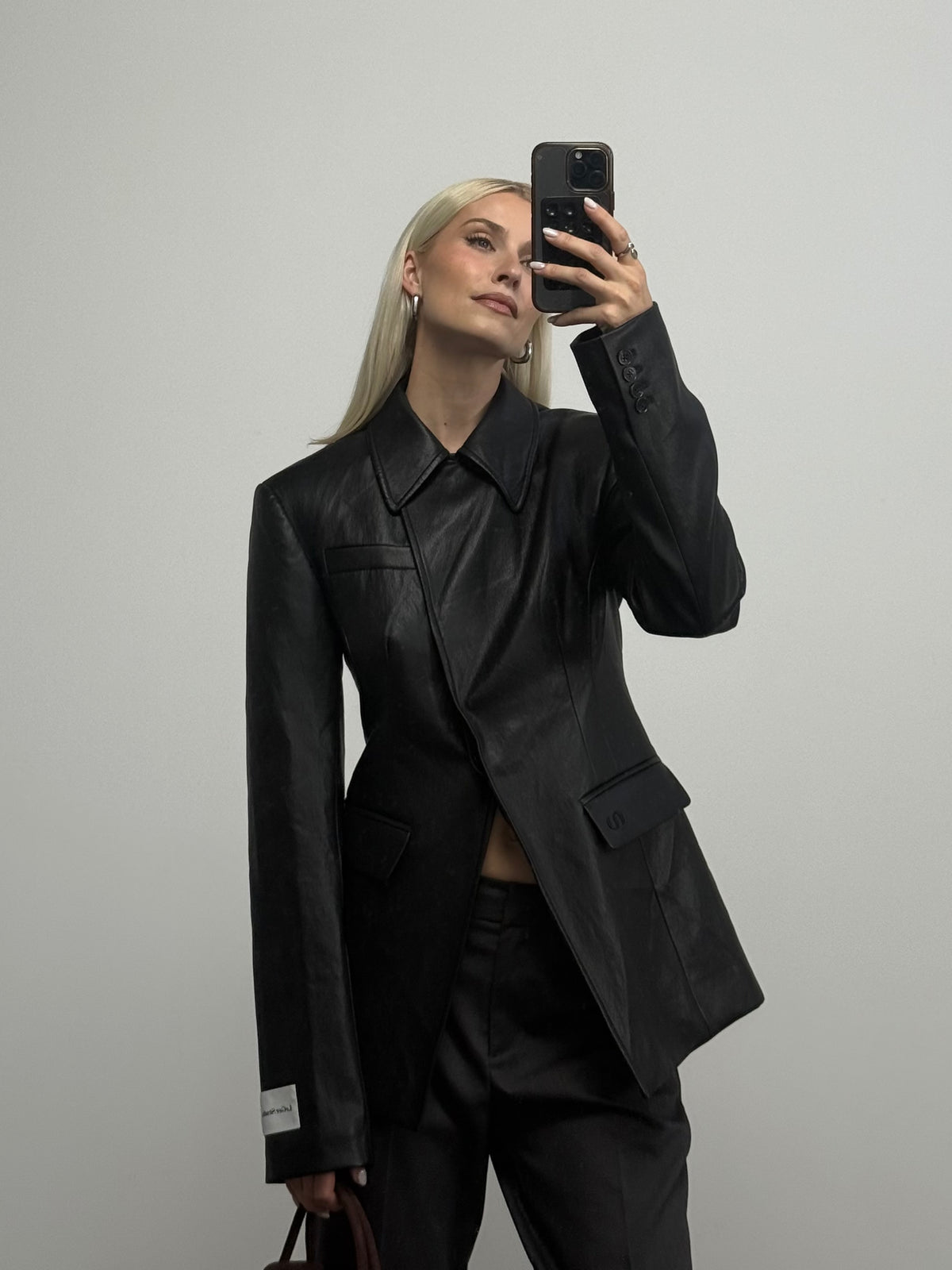 Eine Person mit langen blonden Haaren trägt den schwarzen, taillierten Slim-Fit-Blazer „Orelia“ von LeGer by Lena Gercke in Lederoptik sowie eine schwarze Hose und hält ihr Handy hoch, um vor einem schlichten hellgrauen Hintergrund ein Spiegel-Selfie zu machen.
