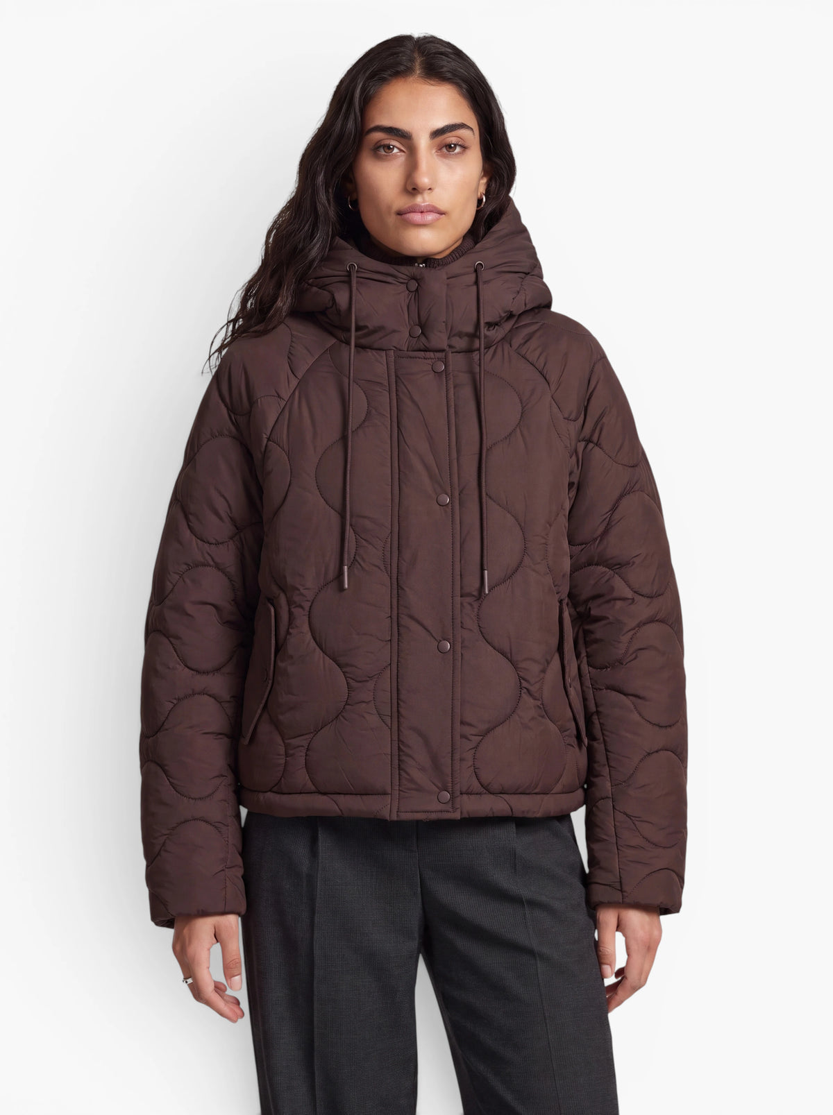 Eine Person trägt die braune „Fray“-Steppjacke im Oversize-Schnitt mit Kapuze, vorne mit Knöpfen und Kordelzügen von LeGer by Lena Gercke, kombiniert mit dunkelgrauen Hosen, und steht vor einem schlichten weißen Hintergrund.