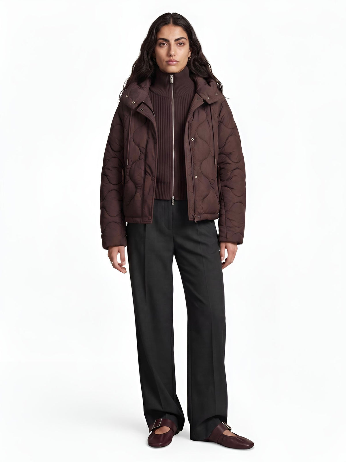 Eine Person steht mit dem Gesicht zur Kamera und trägt die braune „Fray“-Steppjacke im Oversized-Schnitt von LeGer by Lena Gercke, einen gerippten Zip-up-Pullover, dunkelgraue, weite Hosen und braune Loafer vor einem schlichten weißen Hintergrund.