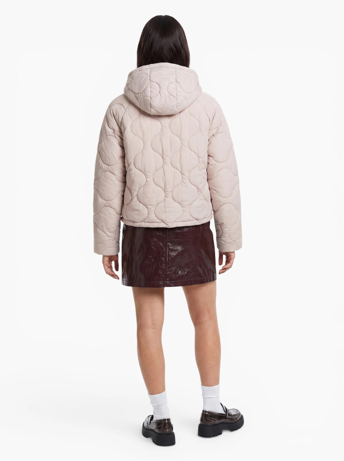 Eine Person steht mit dem Rücken zur Kamera und trägt die gesteppte Kapuzenjacke „Fray“ von LeGer by Lena Gercke, dazu einen kurzen dunklen Rock, weiße Socken und schwarze Schuhe vor einem schlichten weißen Hintergrund.