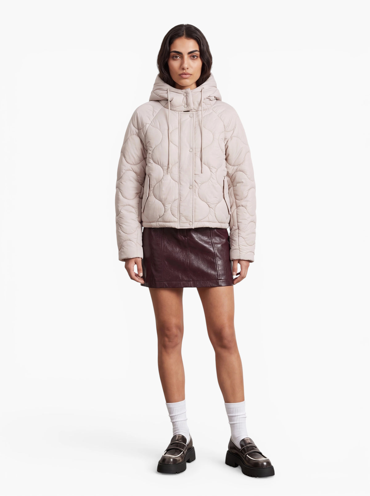 Eine Frau steht mit dem Gesicht nach vorne vor einem schlichten weißen Hintergrund und trägt die Jacke „Fray“ von LeGer by Lena Gercke – eine leichte Steppjacke mit Kapuze im Oversize-Schnitt –, dazu einen kurzen dunklen Lederrock, weiße Socken und schwarze Loafer.