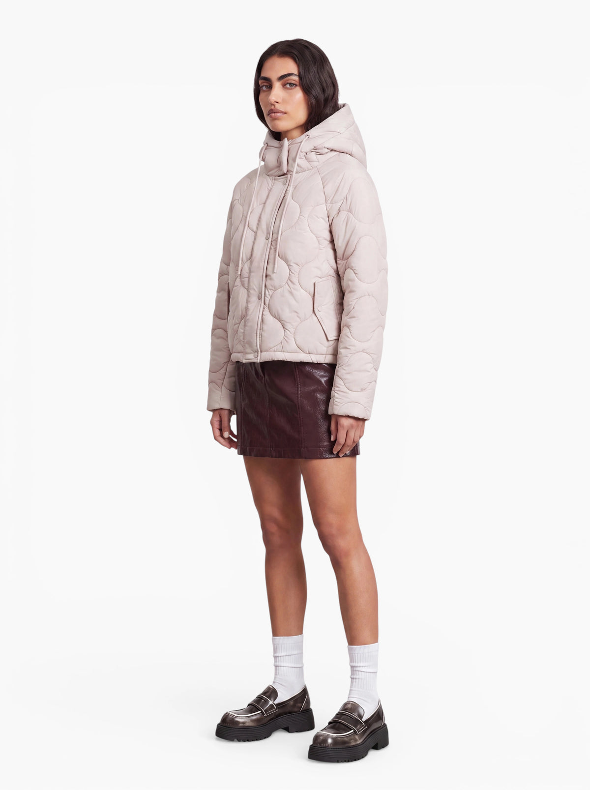 Eine Person, die die „Fray“-Jacke von LeGer by Lena Gercke trägt – eine luftige, gesteppte Steppjacke mit Kapuze im modernen Oversized-Schnitt –, kombiniert mit einem dunklen Minirock, weißen Socken und schwarzen Loafern, steht vor einem schlichten weißen Hintergrund.