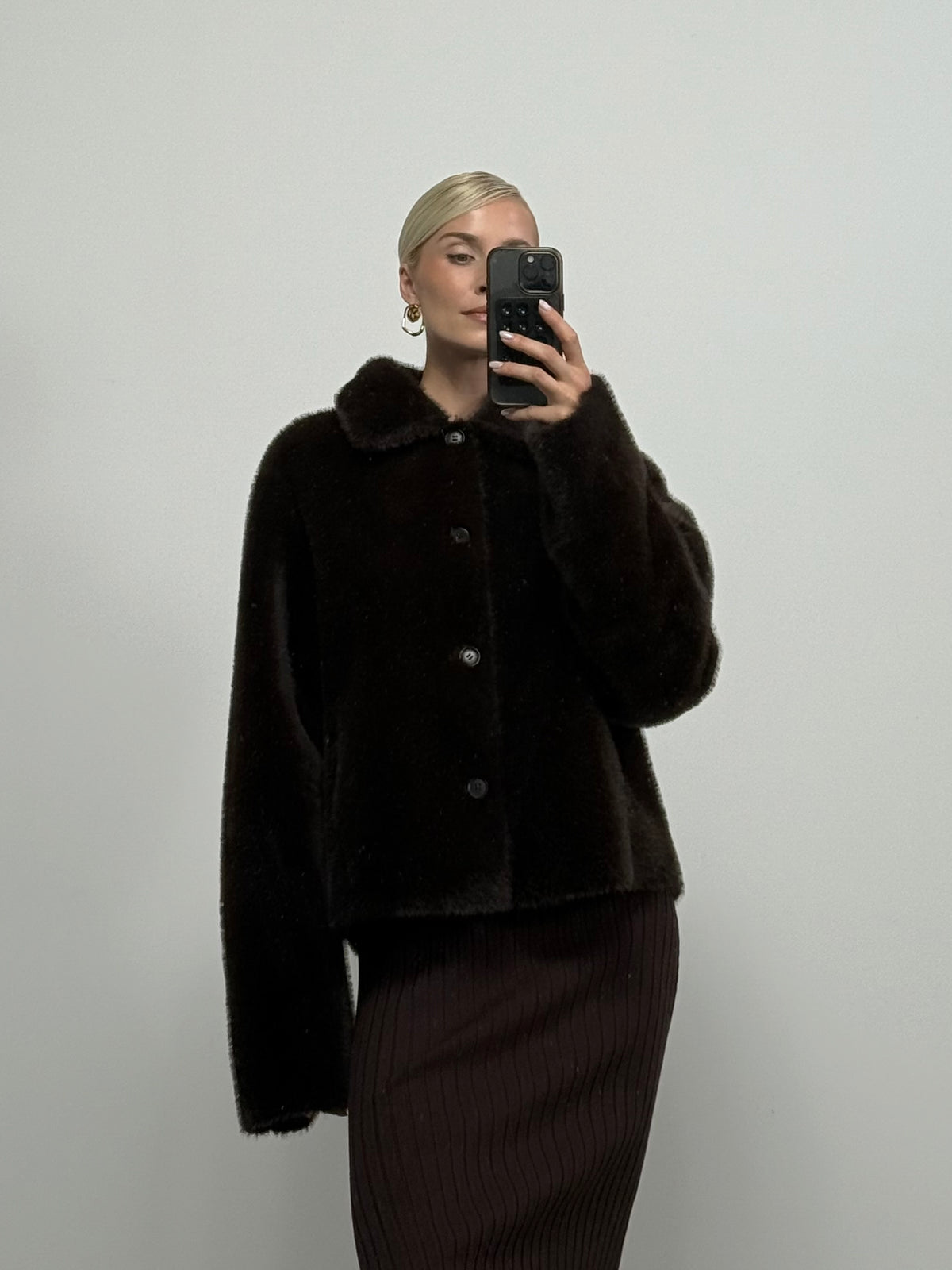 Eine Person macht ein Spiegel-Selfie und trägt die Jacke „Masha“ von LeGer by Lena Gercke in Dunkelbraun mit flauschiger Felloptik sowie einen gerippten dunklen Rock. Sie hat kurzes, nach hinten geglättetes blondes Haar und steht vor einem schlichten hellen Hintergrund.