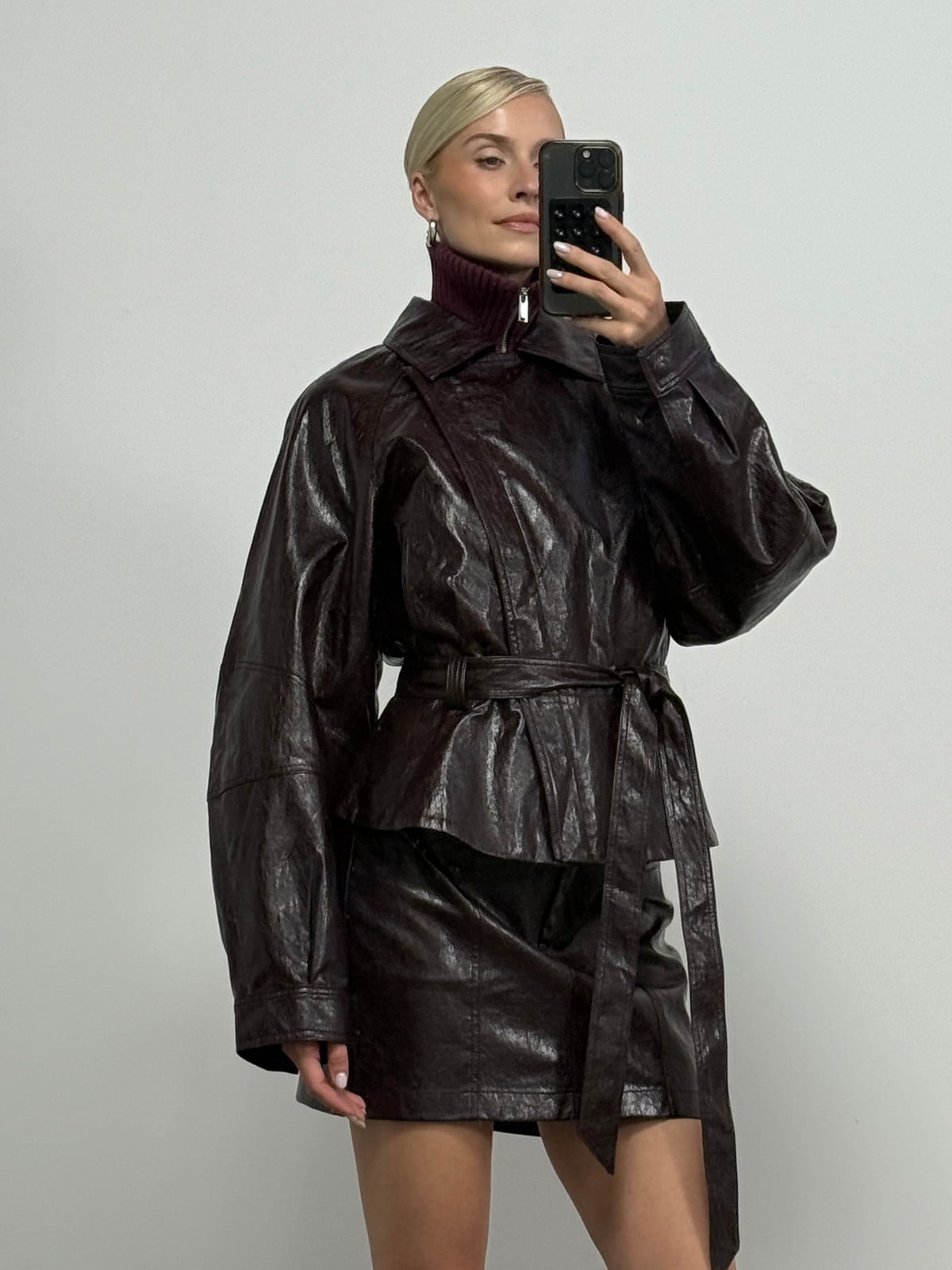Eine Person mit blonden Haaren, die zu einem glatten Dutt hochgesteckt sind, macht ein Spiegel-Selfie. Sie trägt die dunkelbraune „Sina“-Jacke in Lederoptik von LeGer by Lena Gercke mit einem stilvollen Bindegürtel, dazu einen passenden Lederrock und einen weinroten Rollkragenpullover, und steht vor einem schlichten, hellen Hintergrund.