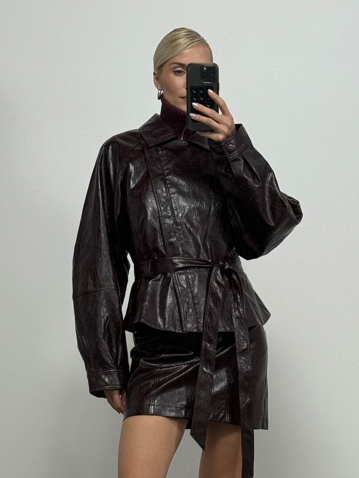 Eine Person in einer dunkelbraunen Kunstlederjacke und dem Rock „Sina“ von LeGer by Lena Gercke posiert vor einem schlichten weißen Hintergrund für ein Spiegel-Selfie. Das Outfit mit seiner Lederoptik und der hohen Taille mit Gürtel wird durch das kurze, nach hinten gegelte blonde Haar der Trägerin abgerundet.