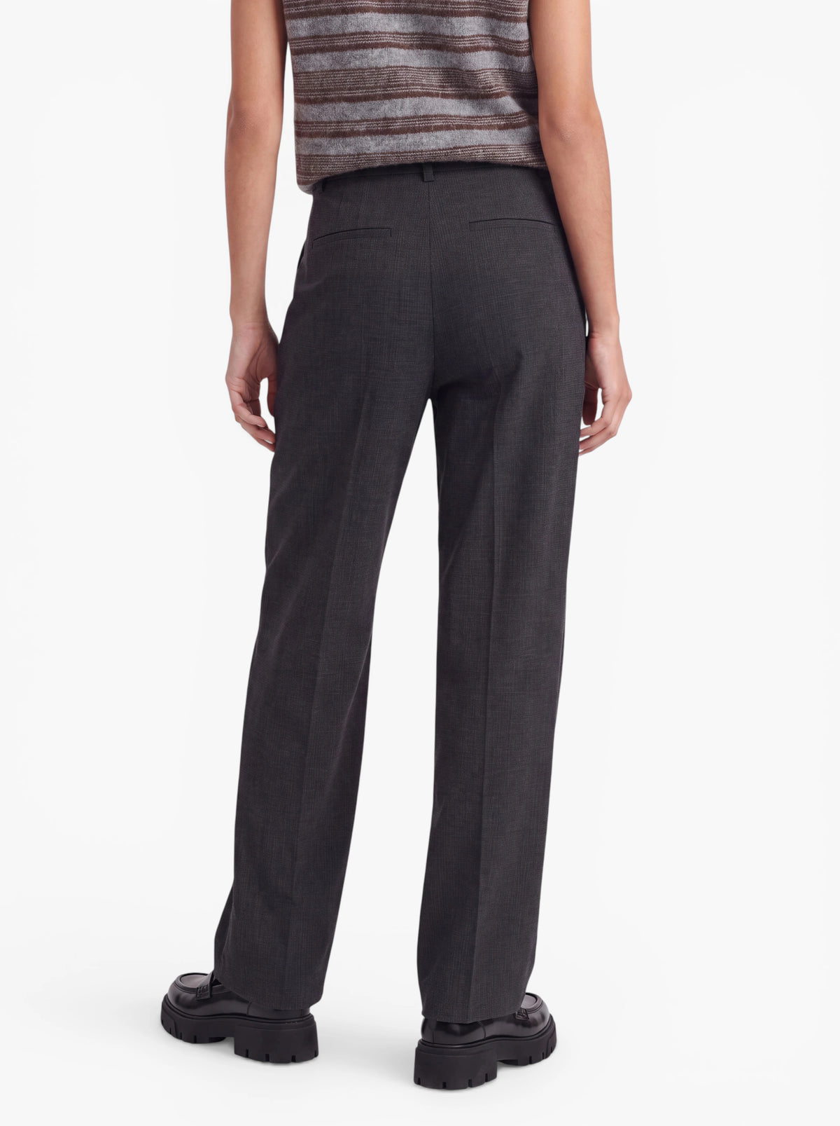 Eine Person, die ein ärmelloses, gestreiftes Oberteil, die dunkelgraue „Julie“-Hose von LeGer by Lena Gercke mit hoher Taille und gepressten Falten sowie schwarze, klobige Schuhe trägt, steht mit dem Rücken zur Kamera und präsentiert die Rückseite ihres Outfits vor einem schlichten weißen Hintergrund.