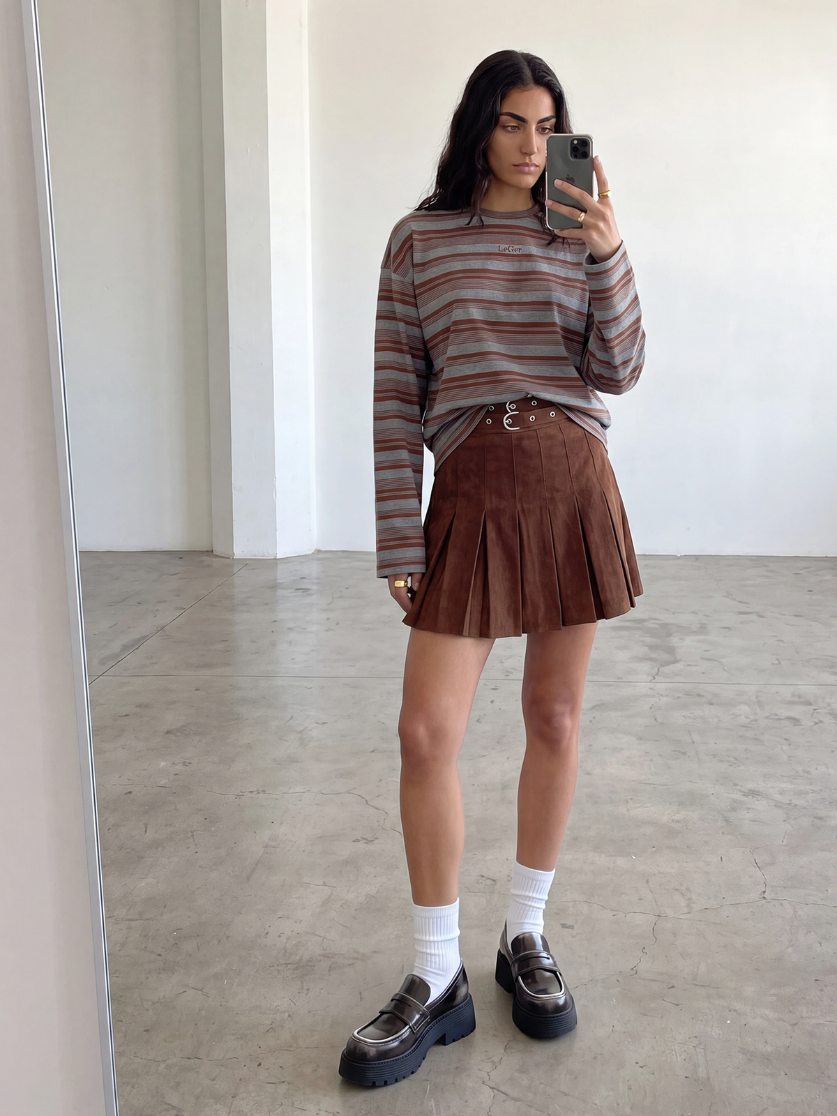 Eine Person, die ein Spiegel-Selfie macht. Sie trägt das gestreifte grau-braune Langarmshirt „Vivian“ von LeGer by Lena Gercke mit gesticktem Logo, dazu einen braunen Faltenrock, weiße Socken und Plateau-Loafer, und steht in einem minimalistisch eingerichteten Raum mit Betonboden und weißen Wänden.