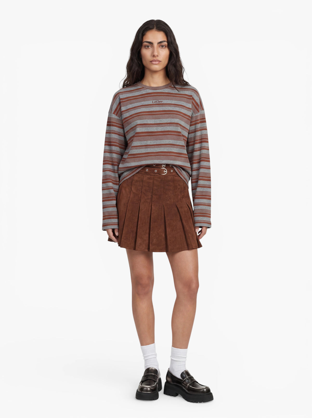 Eine Frau steht mit dem Gesicht zur Kamera und trägt das Langarmshirt „Vivian“ von LeGer by Lena Gercke mit grau-braunen Streifen, dazu einen braunen Faltenrock, weiße Socken und schwarze Plateauschuhe vor einem schlichten weißen Hintergrund.