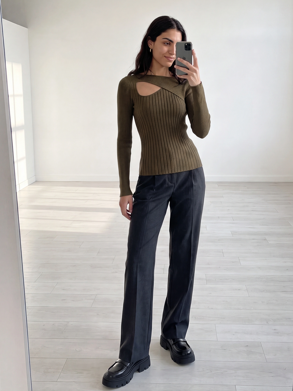 Eine Frau steht in einem minimalistisch gestalteten, lichtdurchfluteten Raum und macht ein Spiegel-Selfie. Sie trägt den Pullover „Selina“ von LeGer by Lena Gercke – einen olivgrünen, gerippten Strickpullover mit asymmetrischem Ausschnitt –, dazu eine dunkelgraue Hose und klobige schwarze Schuhe.