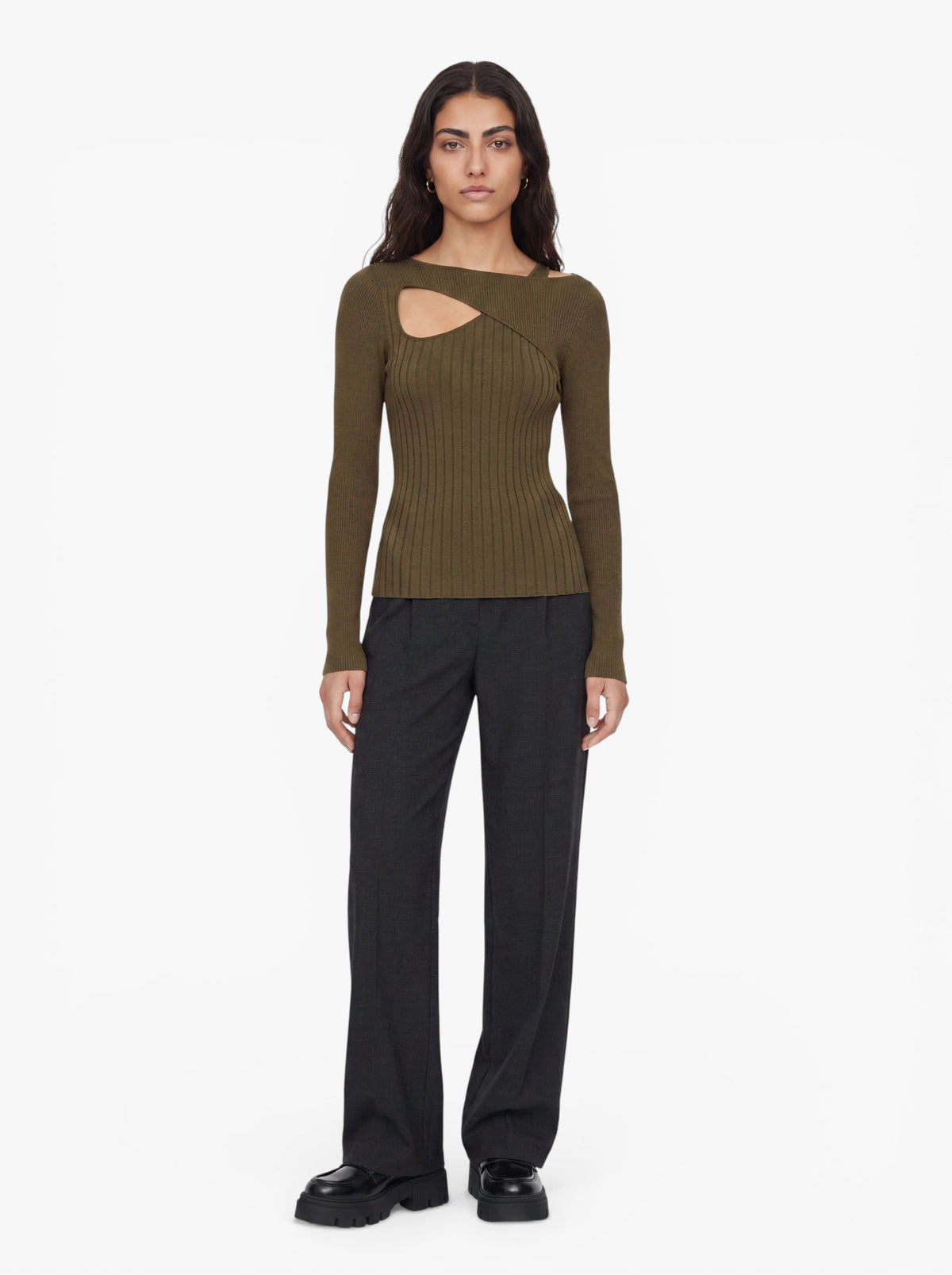 Eine Frau steht mit dem Gesicht nach vorne vor einem schlichten weißen Hintergrund und trägt den olivgrünen „Selina“-Pullover von LeGer by Lena Gercke mit asymmetrischem Ausschnitt und Cut-out im Brustbereich, dazu dunkle, weit geschnittene Hosen und schwarze Plateauschuhe.