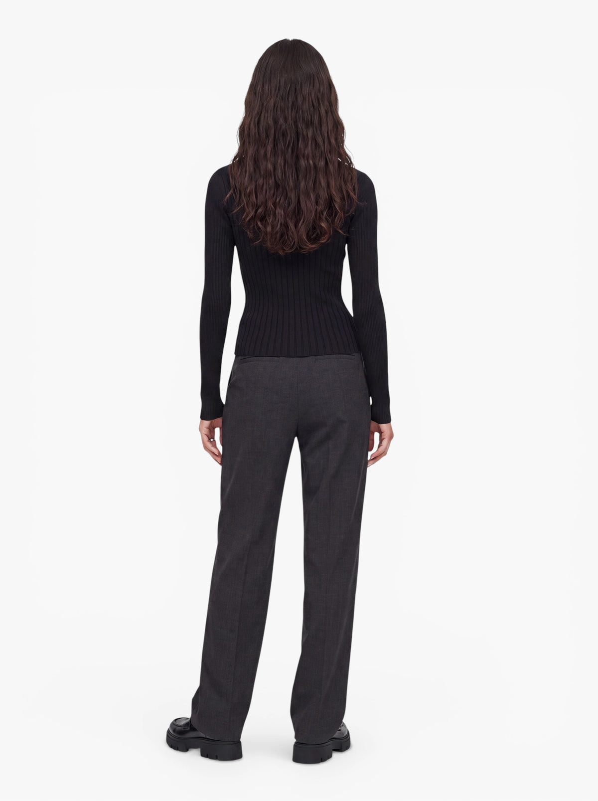 Eine Person mit langen, gewellten Haaren, die den figurbetonten Pullover „Selina“ von LeGer by Lena Gercke mit asymmetrischem Ausschnitt, eine graue Hose und schwarze Schuhe trägt, steht mit dem Rücken zur Kamera vor einem schlichten weißen Hintergrund.