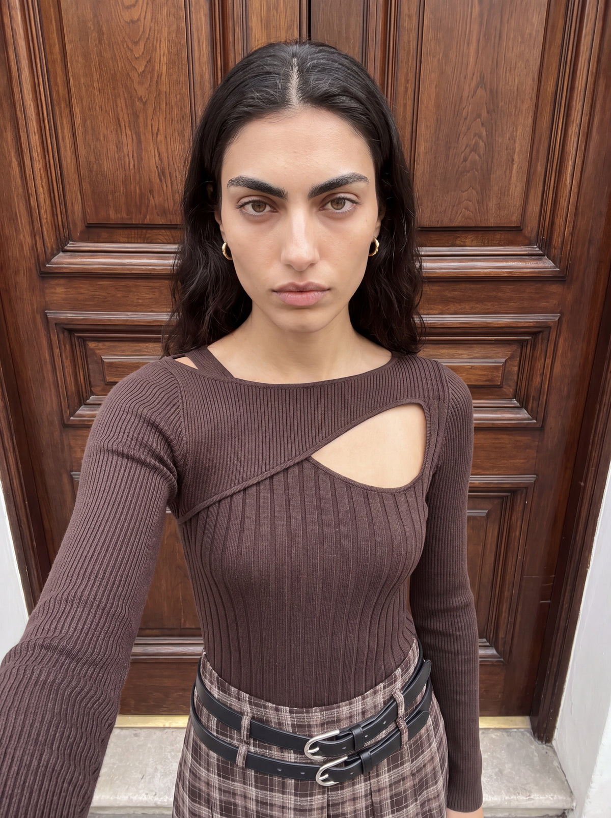 Eine Person mit langen, dunklen Haaren trägt den Pullover „Selina“ von LeGer by Lena Gercke aus olivgrünem Rippstrick mit asymmetrischem Ausschnitt. Dazu trägt sie einen karierten Rock, während sie vor einer Holztür steht und direkt in die Kamera blickt.