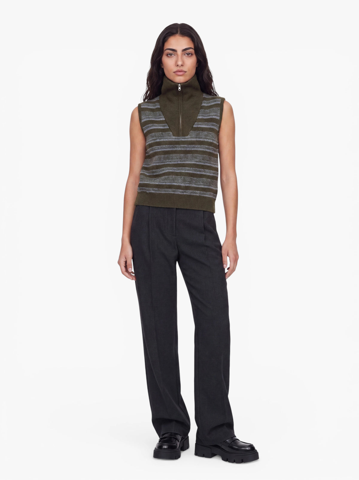 Eine Person mit langen, dunklen Haaren trägt die Weste „Nadia“ aus der LeGer by Lena Gercke-Kollektion mit olivfarbenem Streifenmuster und Reißverschlusskragen, dazu eine dunkle Hose mit weitem Bein und schwarze Schuhe, vor einem schlichten weißen Hintergrund.