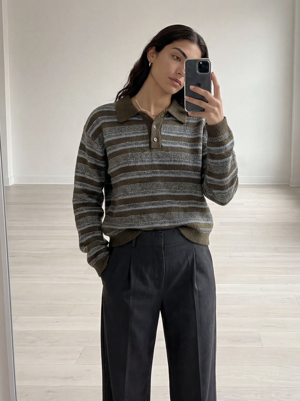 Eine Person steht in einem minimalistischen, lichtdurchfluteten Raum mit hellem Holzboden und macht ein Spiegel-Selfie. Sie trägt den Pullover „Delia“ von LeGer by Lena Gercke mit olivgrünen Streifen und einem klassischen Hemdkragen, dazu eine dunkle Hose mit hoher Taille.