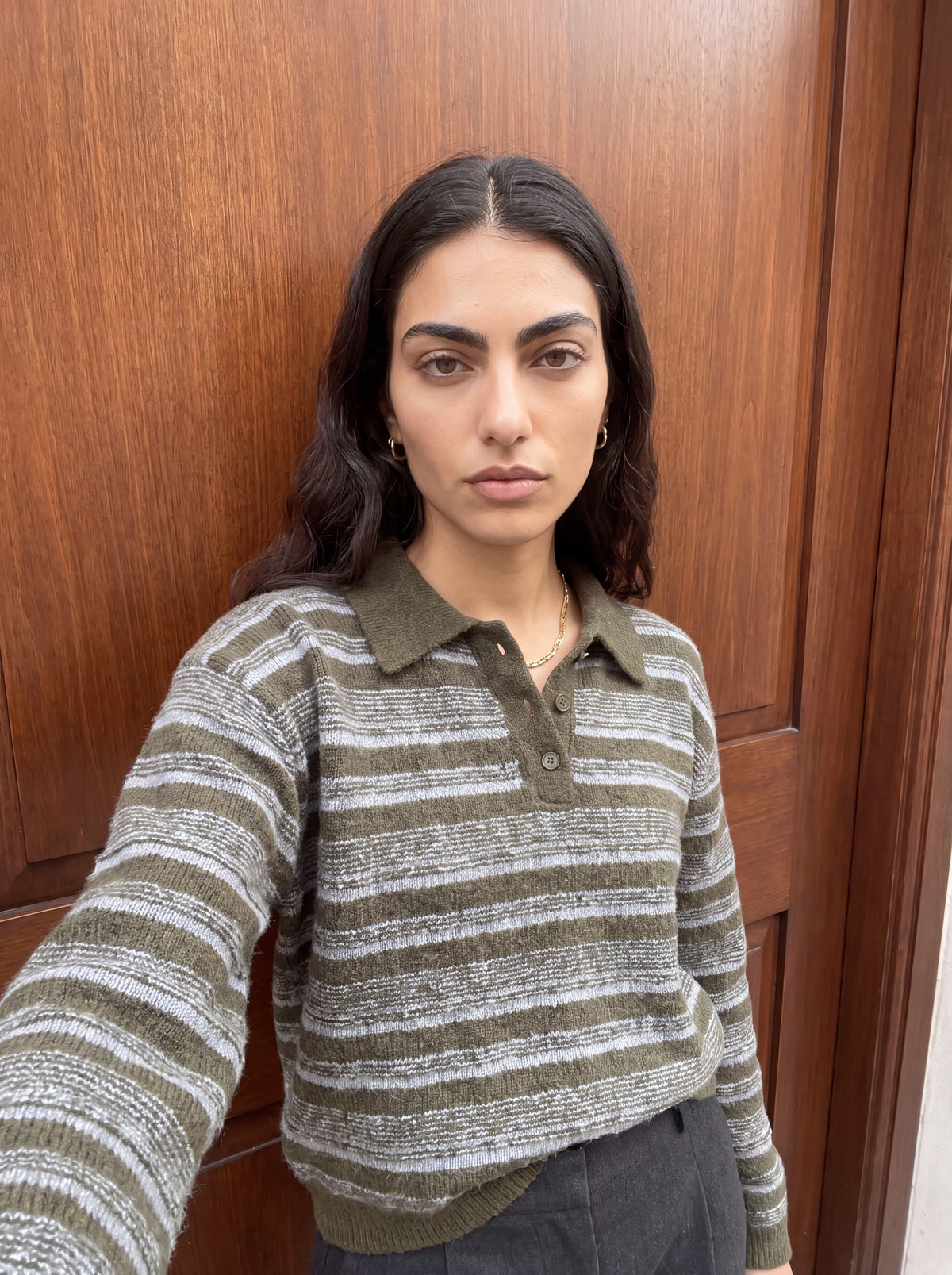 Eine Frau mit langen dunklen Haaren, die den Pullover „Delia“ von LeGer by Lena Gercke mit olivgrünen Streifen und einem Hemdkragen trägt, steht vor einer Holztür und macht mit ausdruckslosem Gesicht ein Selfie.