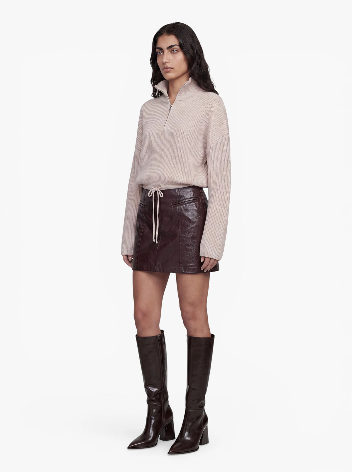 Eine Person trägt den „Michelle“-Pullover von LeGer by Lena Gercke – einen beigen, kurzen Strickpullover mit Rippenstruktur und hohem Kragen – kombiniert mit einem dunkelbraunen Lederminirock mit Kordelzug und kniehohen braunen Stiefeln. Der Hintergrund ist schlicht weiß.