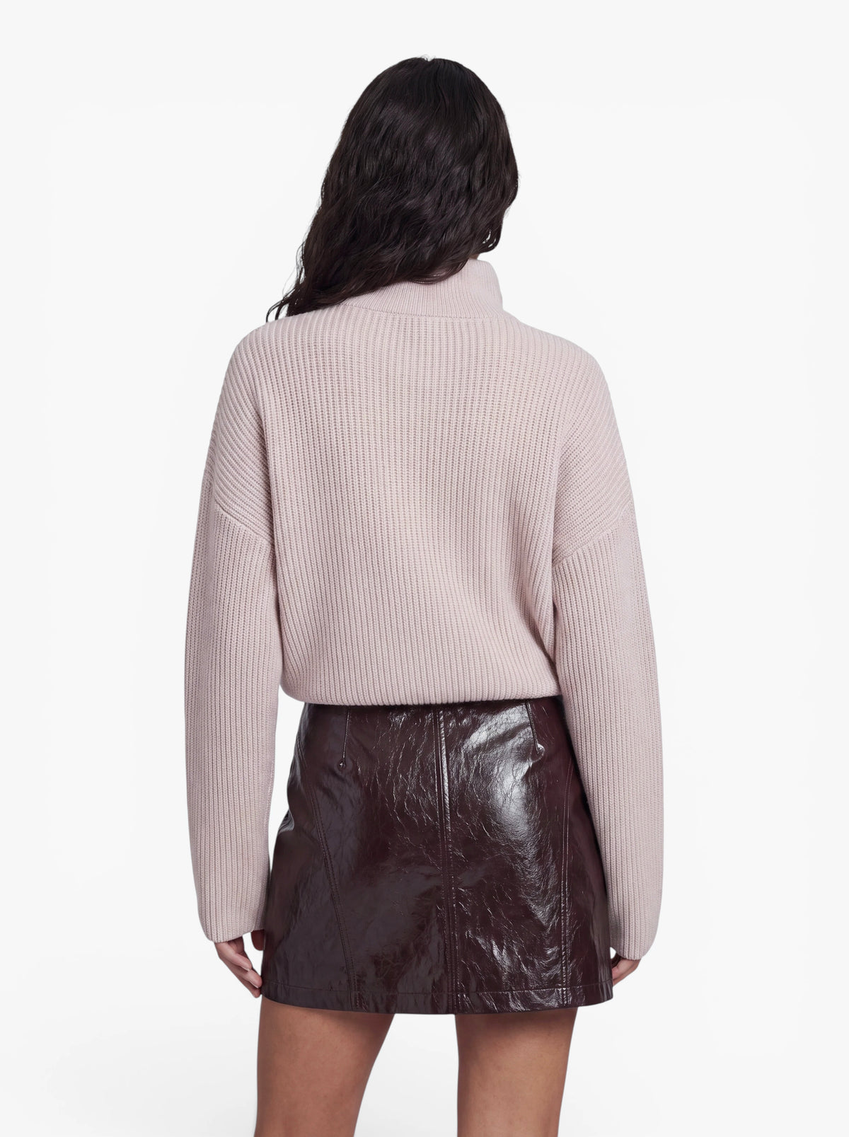 Eine Person mit langen, dunklen Haaren, die den Pullover „Michelle“ von LeGer by Lena Gercke in Hellrosa und einen glänzenden, dunkelbraunen Lederminirock trägt, steht mit dem Rücken zur Kamera vor einem schlichten weißen Hintergrund.
