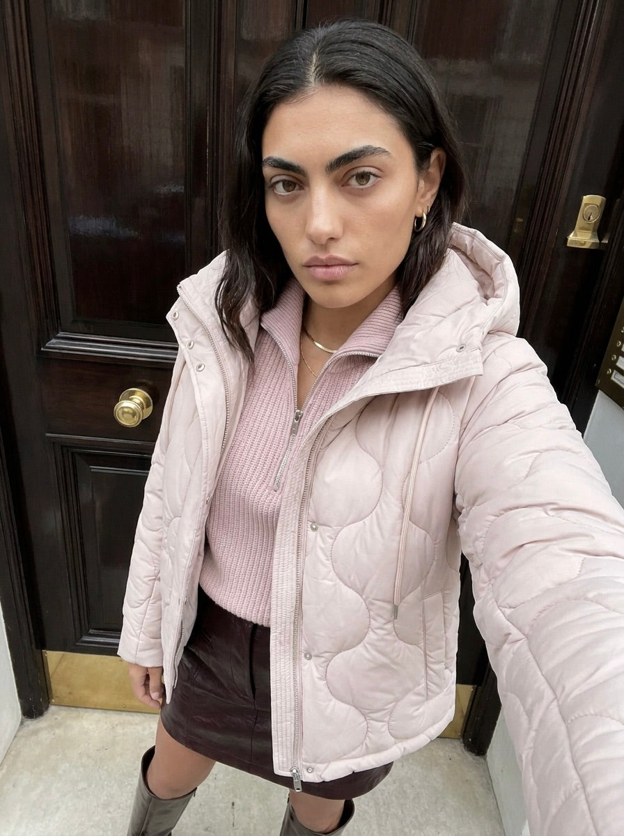 Eine Frau mit langen dunklen Haaren, gekleidet in eine hellrosa Steppjacke mit hohem Kragen und den Pullover „Michelle“ von LeGer by Lena Gercke – einen rosa, fein gerippten, kurzen Strickpullover –, dazu einen dunklen Rock, steht vor einer Holztür und macht ein Selfie.