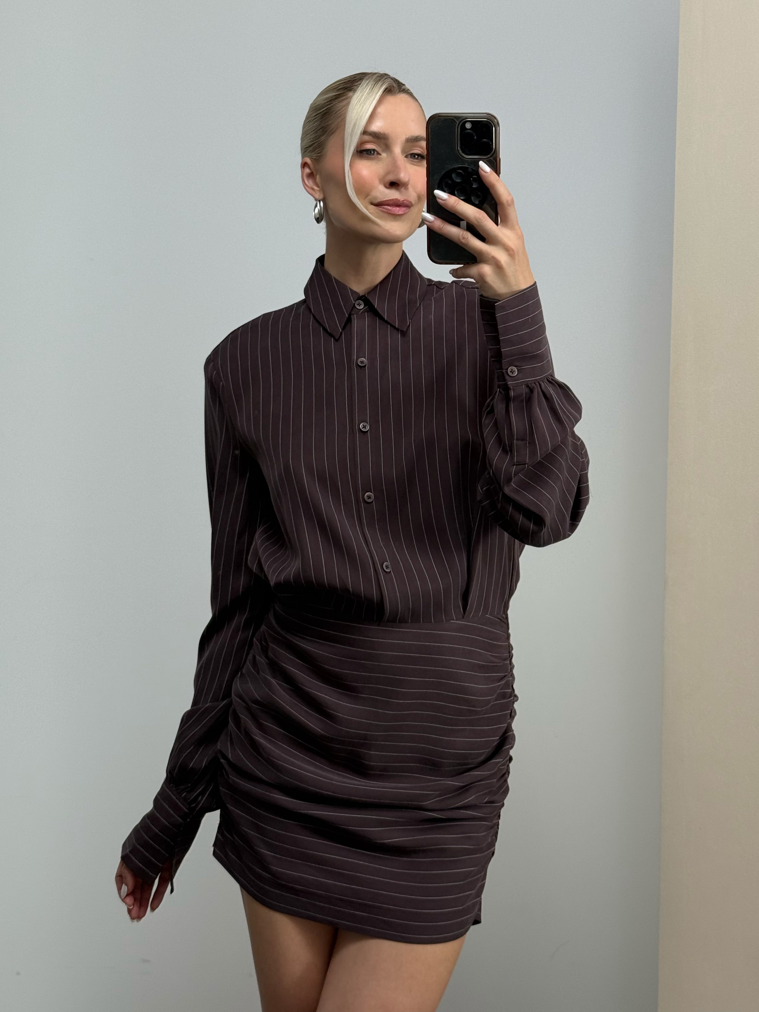 Eine Frau posiert für ein Spiegel-Selfie und trägt dabei das Kleid „Madita“ von LeGer by Lena Gercke, das aus einem dunklen, gestreiften Hemd mit Knöpfen und einem dazu passenden, gestreiften Rock mit Rüschenverzierungen besteht.